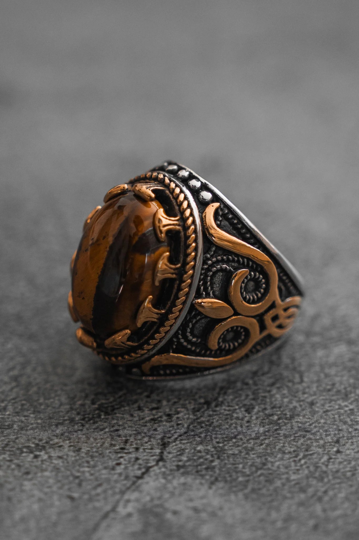 The Royal Revenge Ring by Relentless Betrayal is a handcrafted heirloom featuring an oval brown gemstone set in a gold and silver band with intricate swirling patterns, elegantly showcased against a textured gray background.