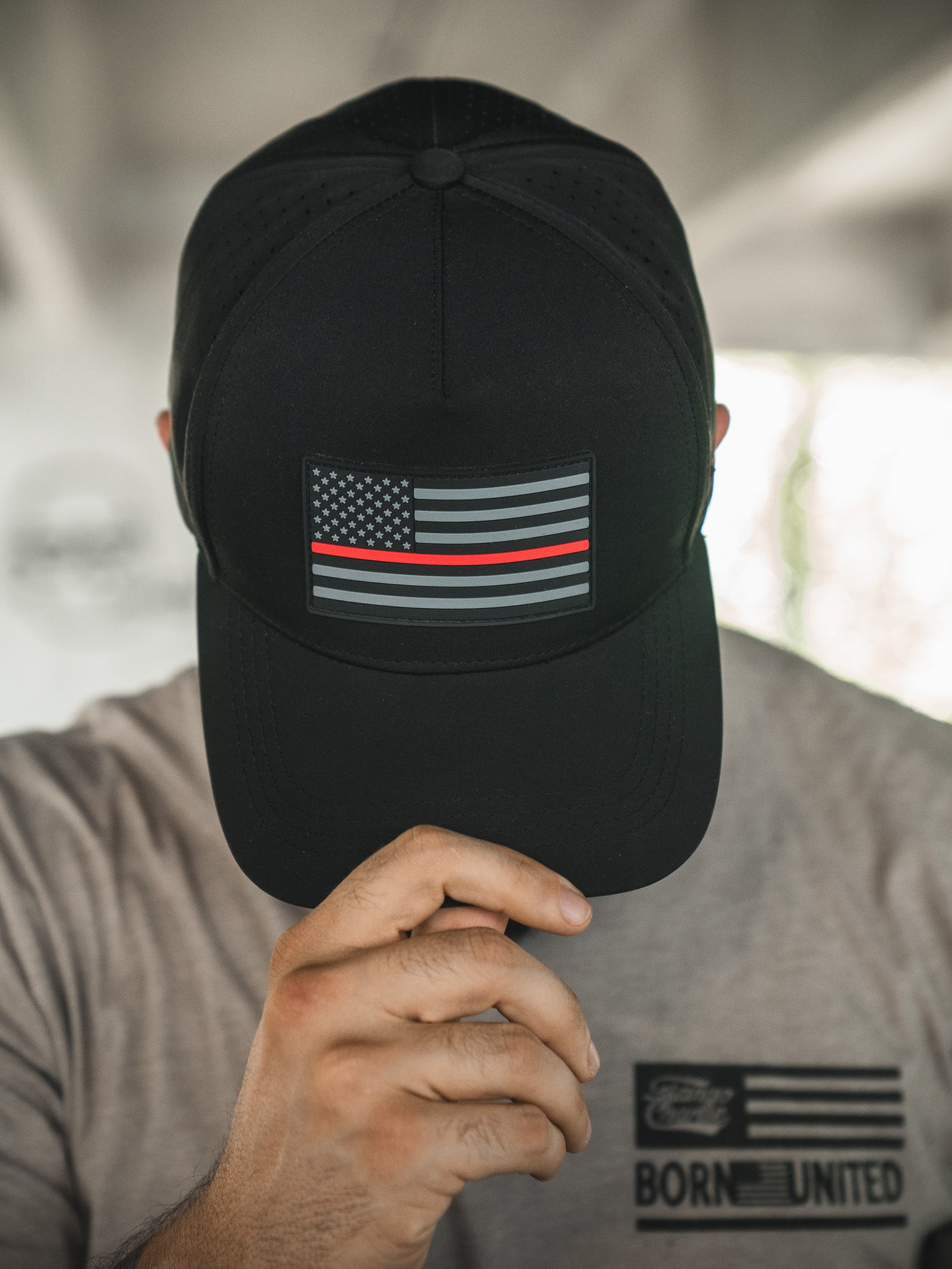 A person wearing the Born United Premium Flag Snapback - TRL by Born United Private Label, featuring a subdued American flag with a thin red line, partially covering their face, and a gray shirt with "Born United" printed on it.