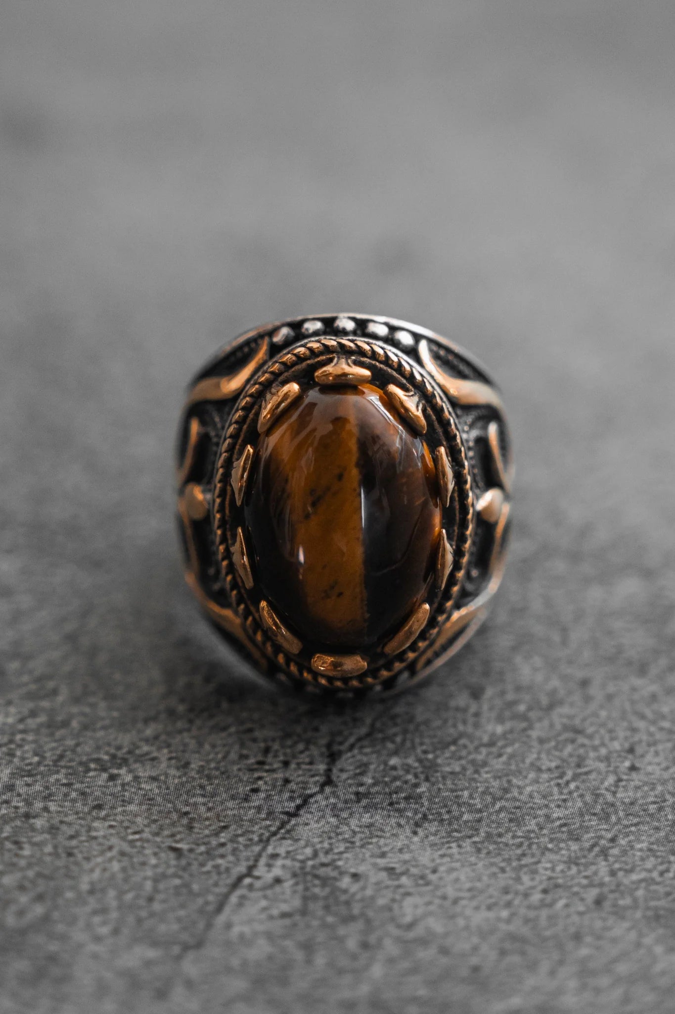 Handcrafted by Relentless Betrayal, the Royal Revenge Ring is an heirloom piece featuring an oval brown gemstone on an ornate metallic band with intricate patterns. Its textured gray background amplifies its vintage charm.