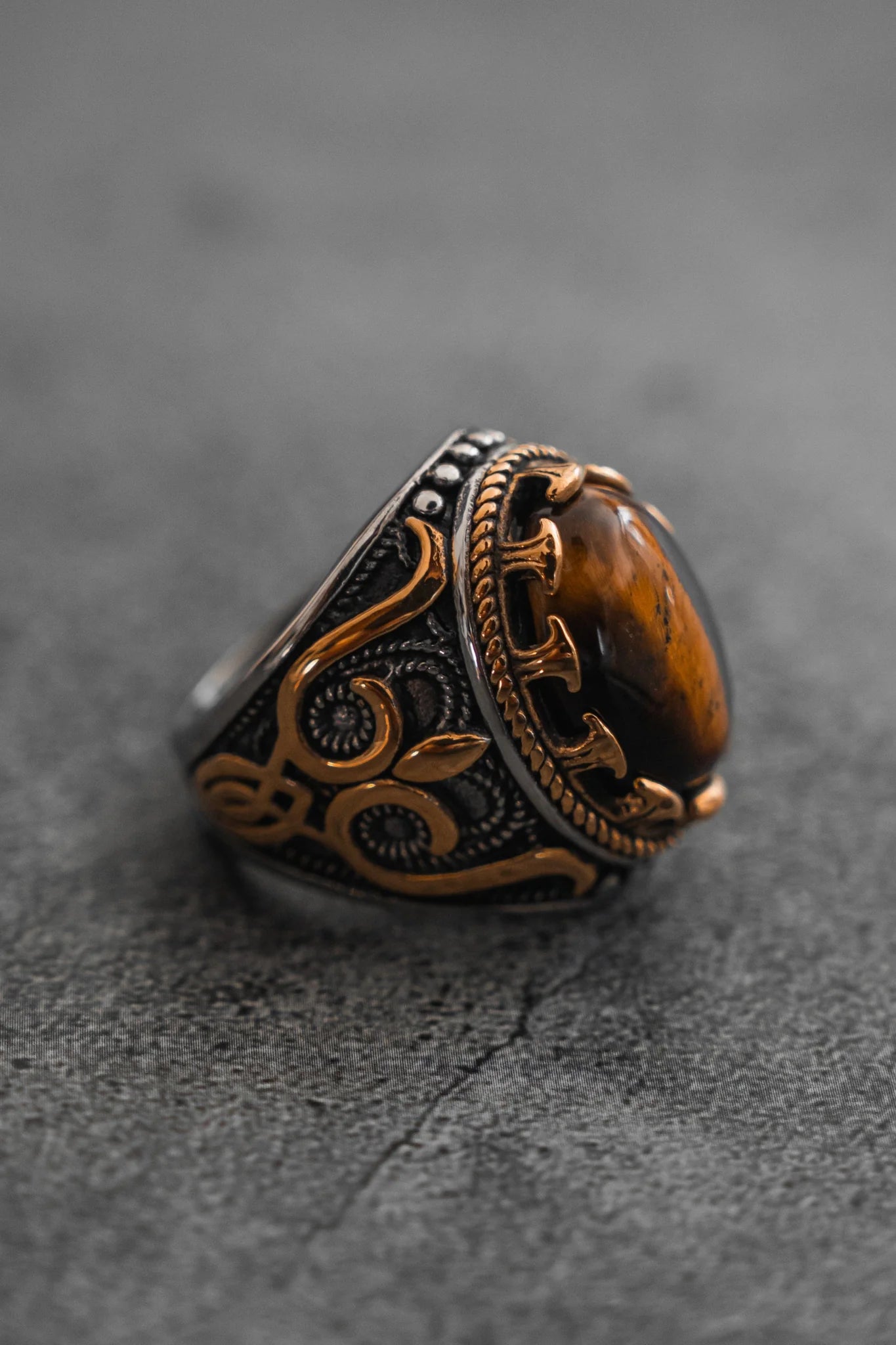 Placed on a textured gray surface, the Royal Revenge Ring by Relentless Betrayal features intricate golden floral designs with a large oval amber stone set in a dark metal band, making it a handcrafted heirloom fit for royalty.