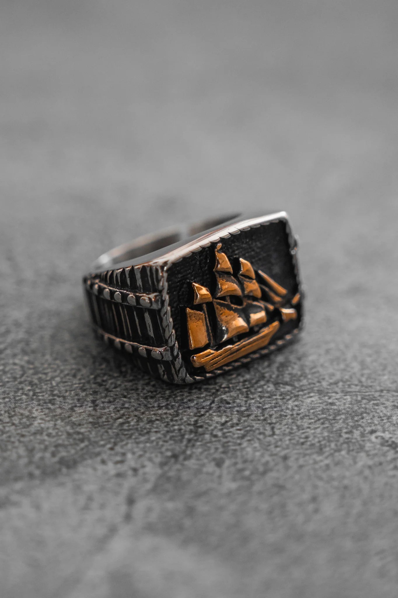 The Queen Annes Revenge Ring by Relentless Betrayal is a silver piece with an engraved ship set against a dark backdrop on a textured gray surface, evoking pirate jewelry.
