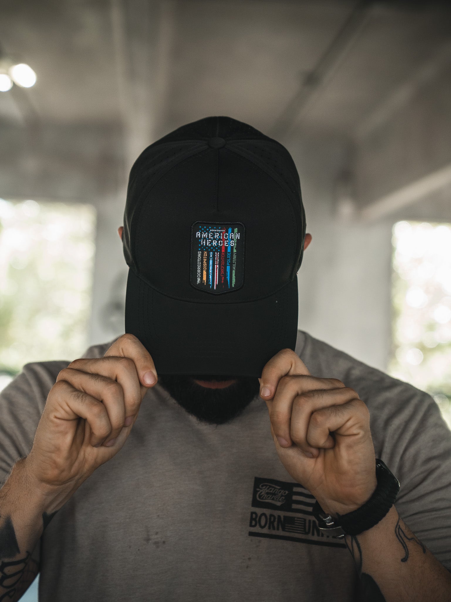 A person in a light gray t-shirt and black cap holds the brim, facing down. The Born United Private Label cap features a small patch with multicolored lines and text reading "American Heroes" from the Born United Premium Snapback line.