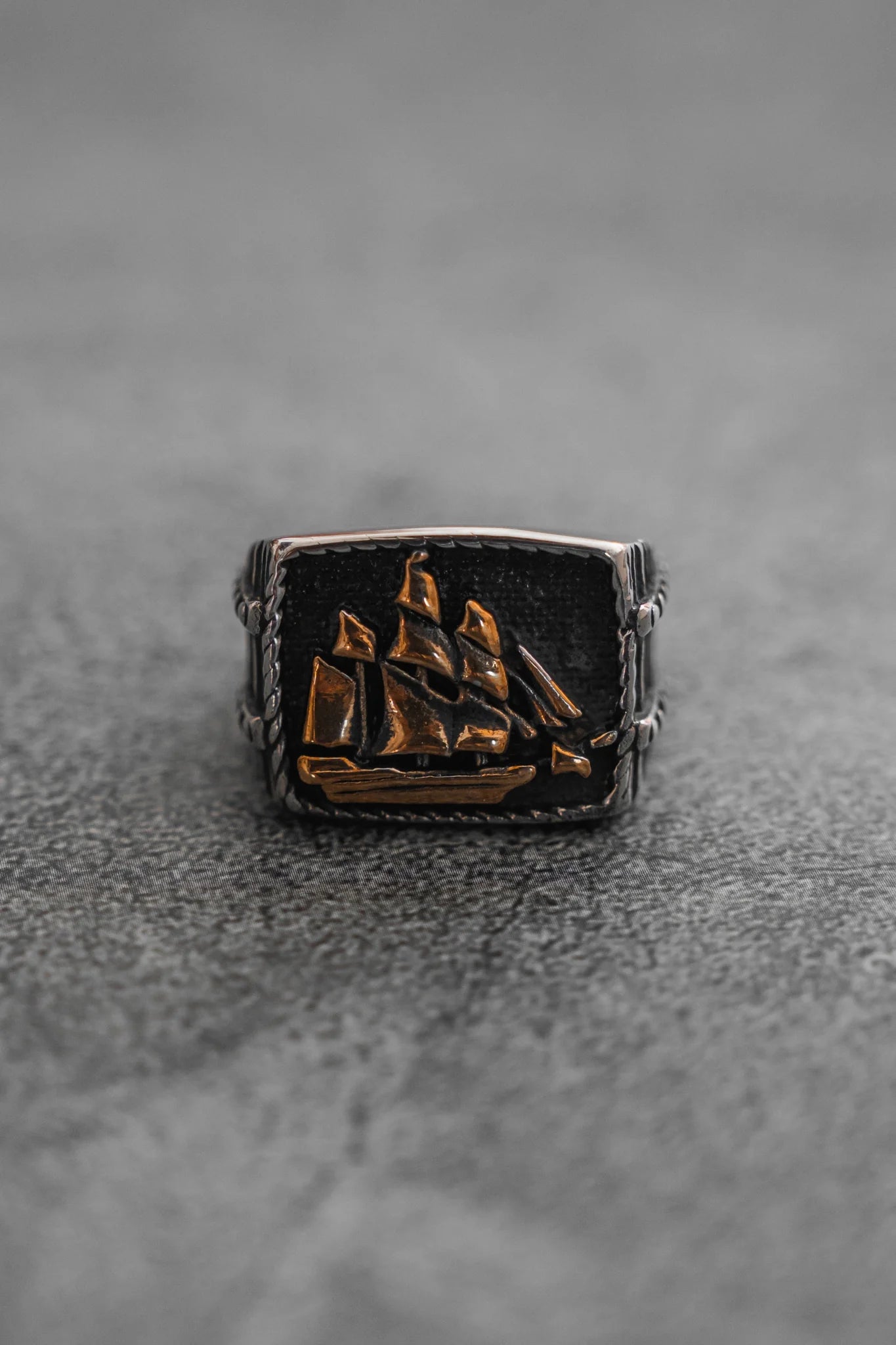 The Queen Annes Revenge Ring by Relentless Betrayal is a silver piece inspired by pirate jewelry. It features an embossed sailing ship on a textured dark background, capturing maritime allure and intricate details reminiscent of treasures like Blackbeards. Displayed on a gray surface.