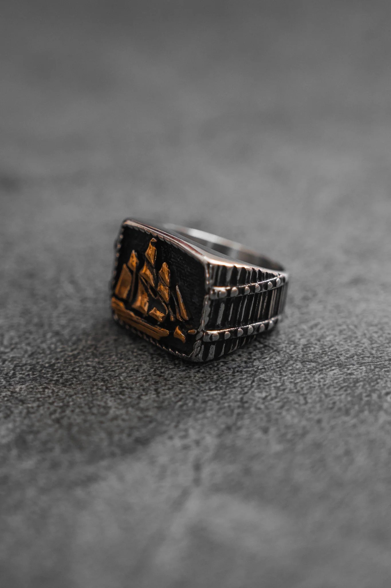 The Queen Annes Revenge Ring by Relentless Betrayal is a silver ring intricately detailed with an engraved ship design, capturing the allure of pirate jewelry, elegantly resting on a textured gray surface inspired by Blackbeards famous voyages.