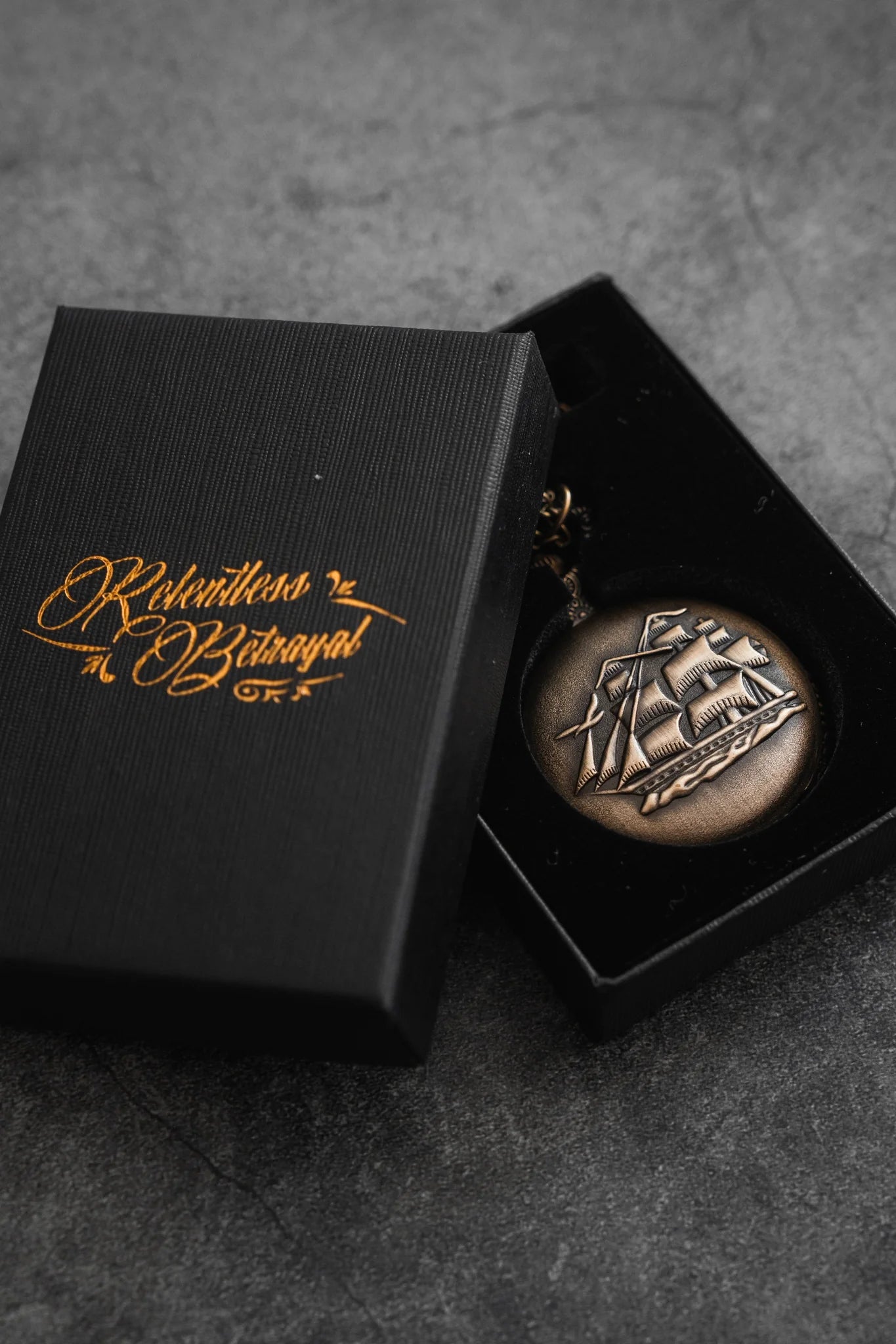 A bronze-colored medal with a pirate ship design is displayed in a black box adorned with gold script reading The Legacy Pocket Watch by Relentless Betrayal, set against a textured gray surface hinting at seafaring adventures and hidden treasures.