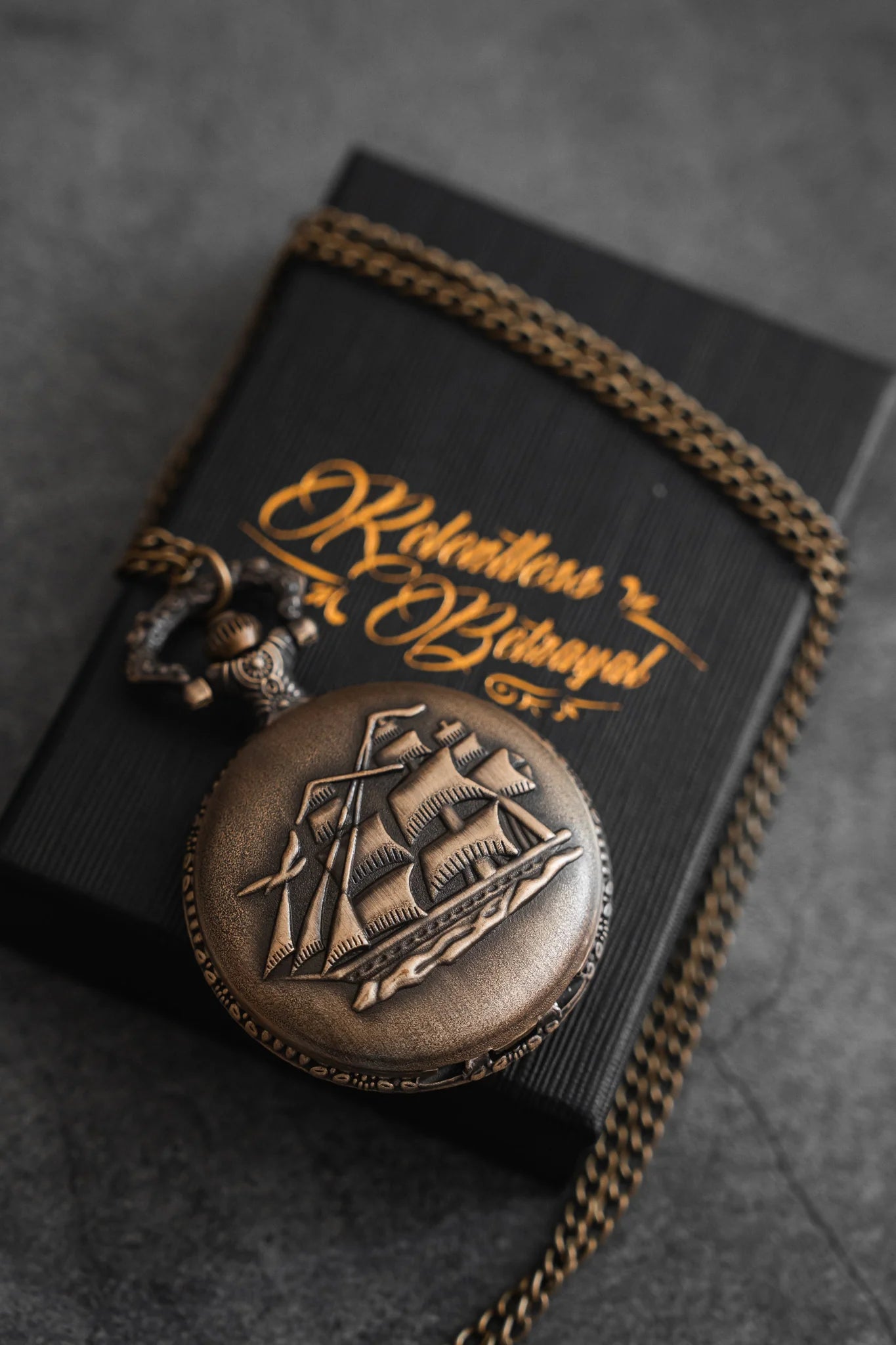 The Legacy Pocket Watch by Relentless Betrayal features a pirate ship engraving and rests on a textured black box with cursive gold writing. A bronze chain with an anchor charm partially wraps the box, all placed against a dark gray background.