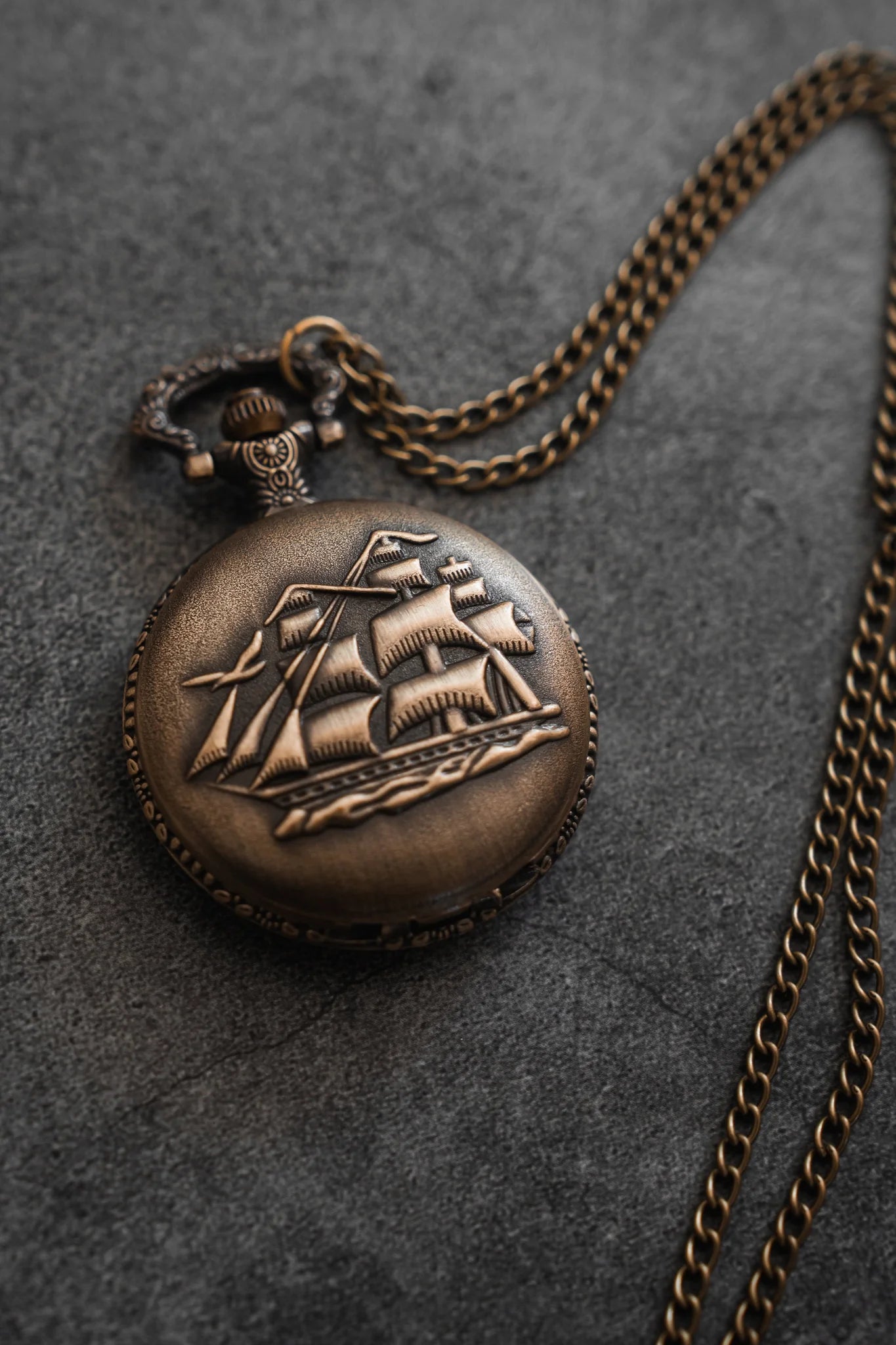 The Legacy Pocket Watch by Relentless Betrayal features an ornate pirate ship engraving on its antique brass cover, resting on a grey stone surface. It comes with a long, intricate chain attached to the top.