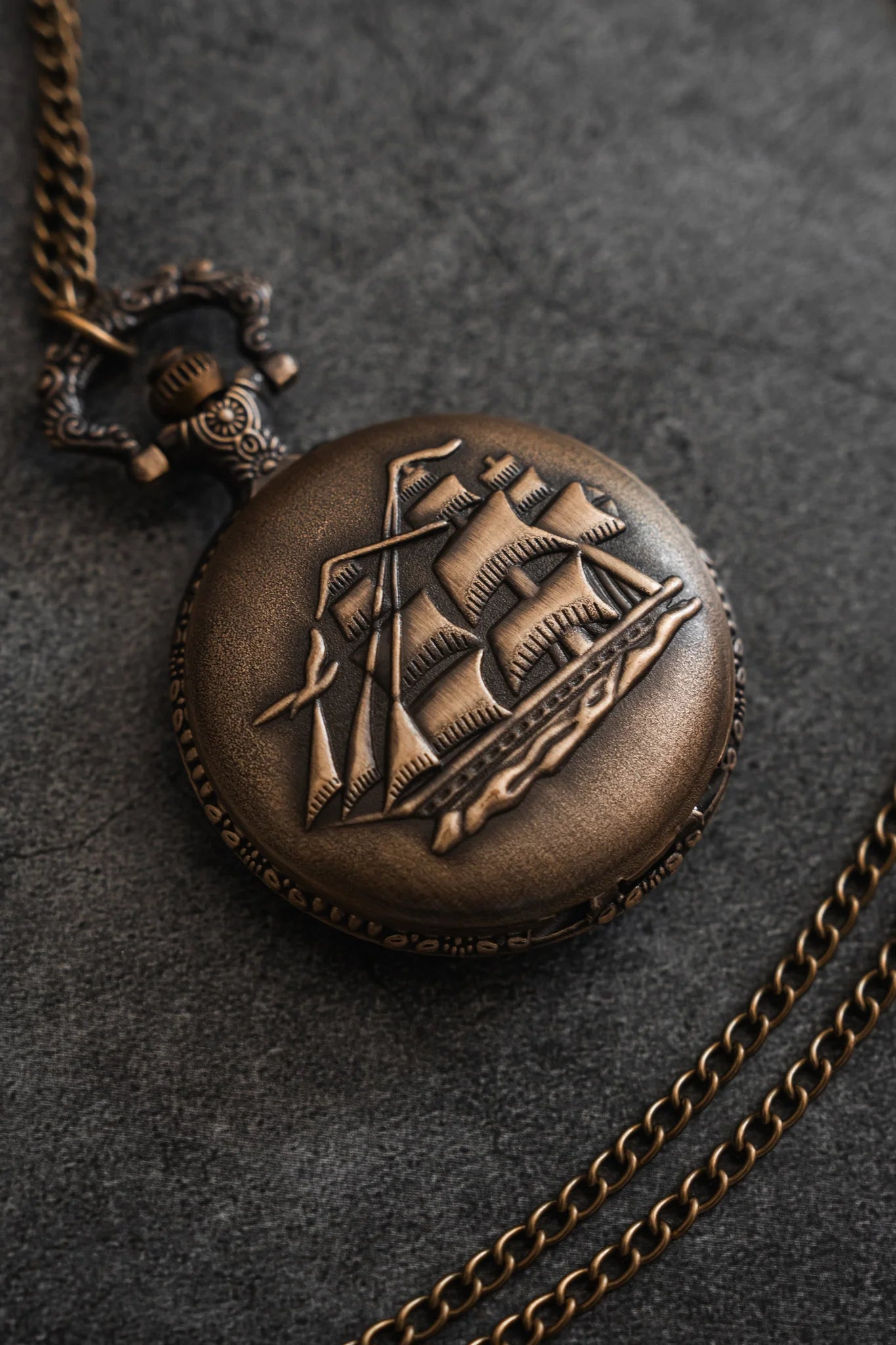 The Legacy Pocket Watch by Relentless Betrayal is a vintage brass pocket watch with a chain, showcasing an embossed pirate ship design on the cover, elegantly displayed on a textured dark surface.