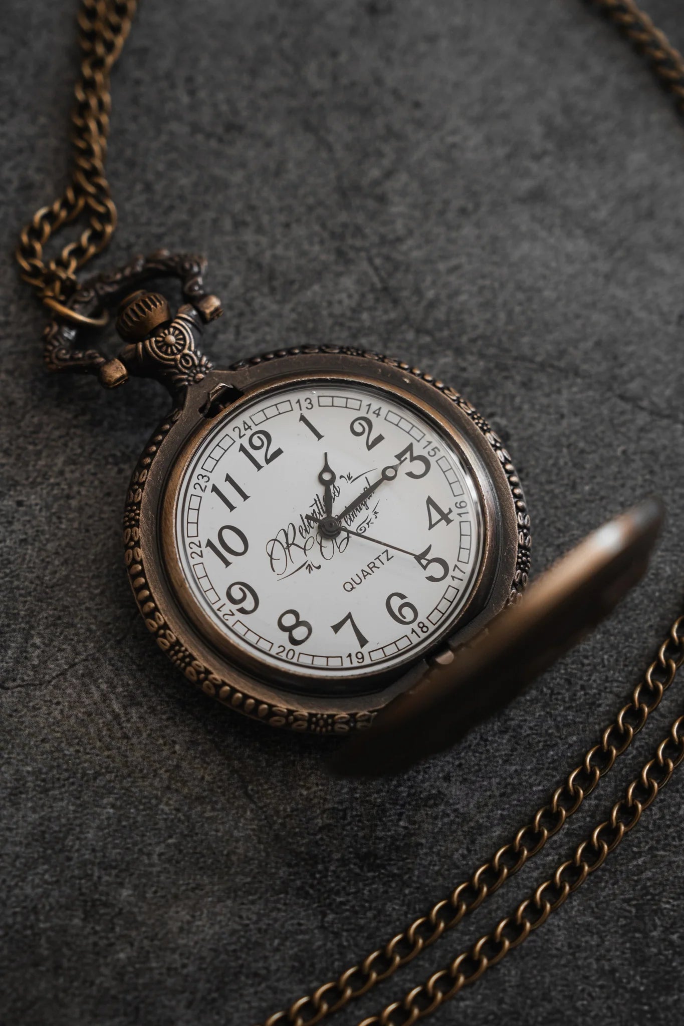 The Legacy Pocket Watch by Relentless Betrayal features an ornate, antique design with a bronze finish and a white face with black numerals, resting on a textured gray surface. Its intricate chain evokes memories of pirate treasure.