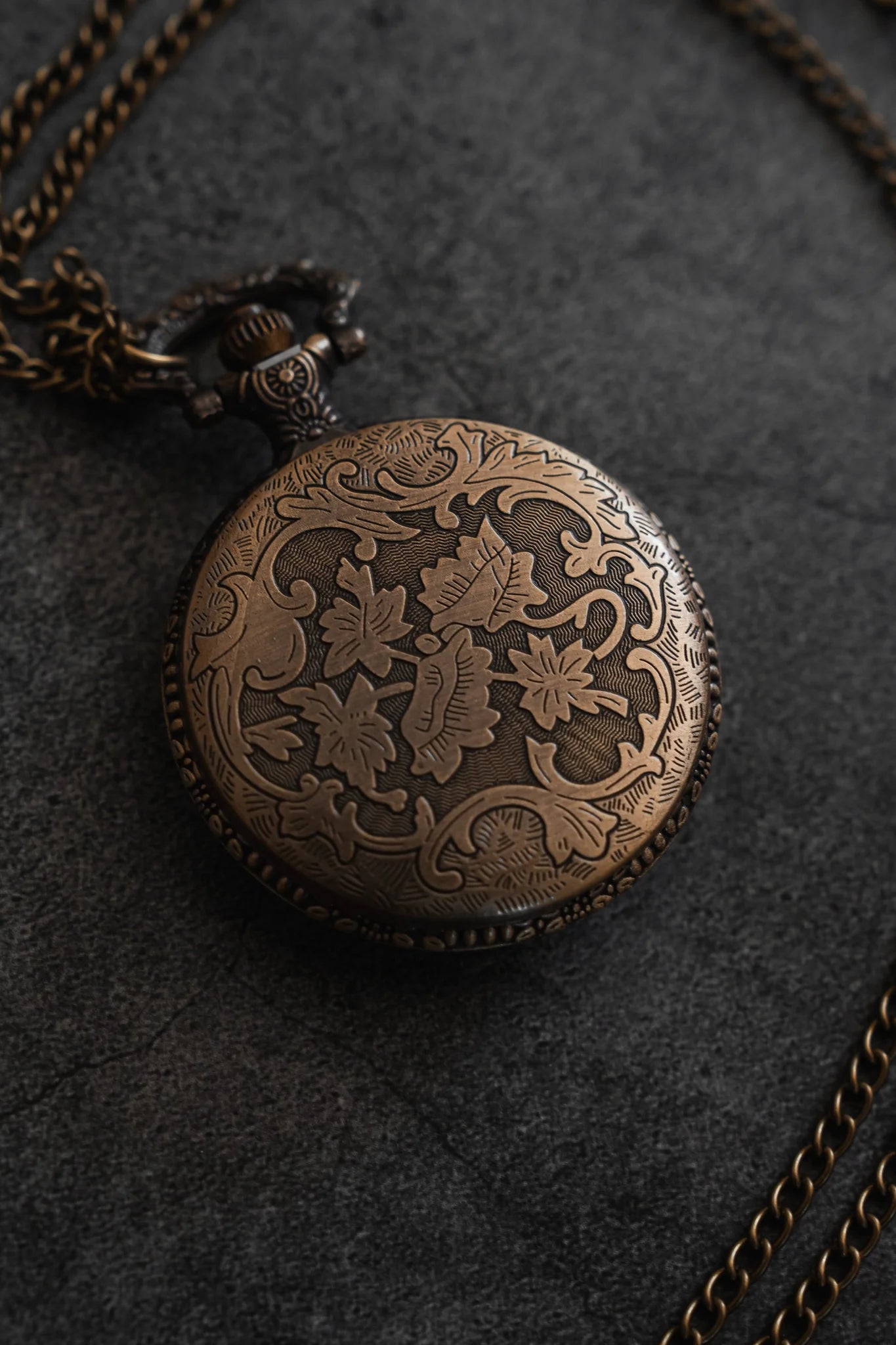 Resting closed on a gray textured surface, The Legacy Pocket Watch by Relentless Betrayal features intricate floral and leaf engravings. A chain, reminiscent of a pirate ship anchor, is attached and elegantly curls around the watch.