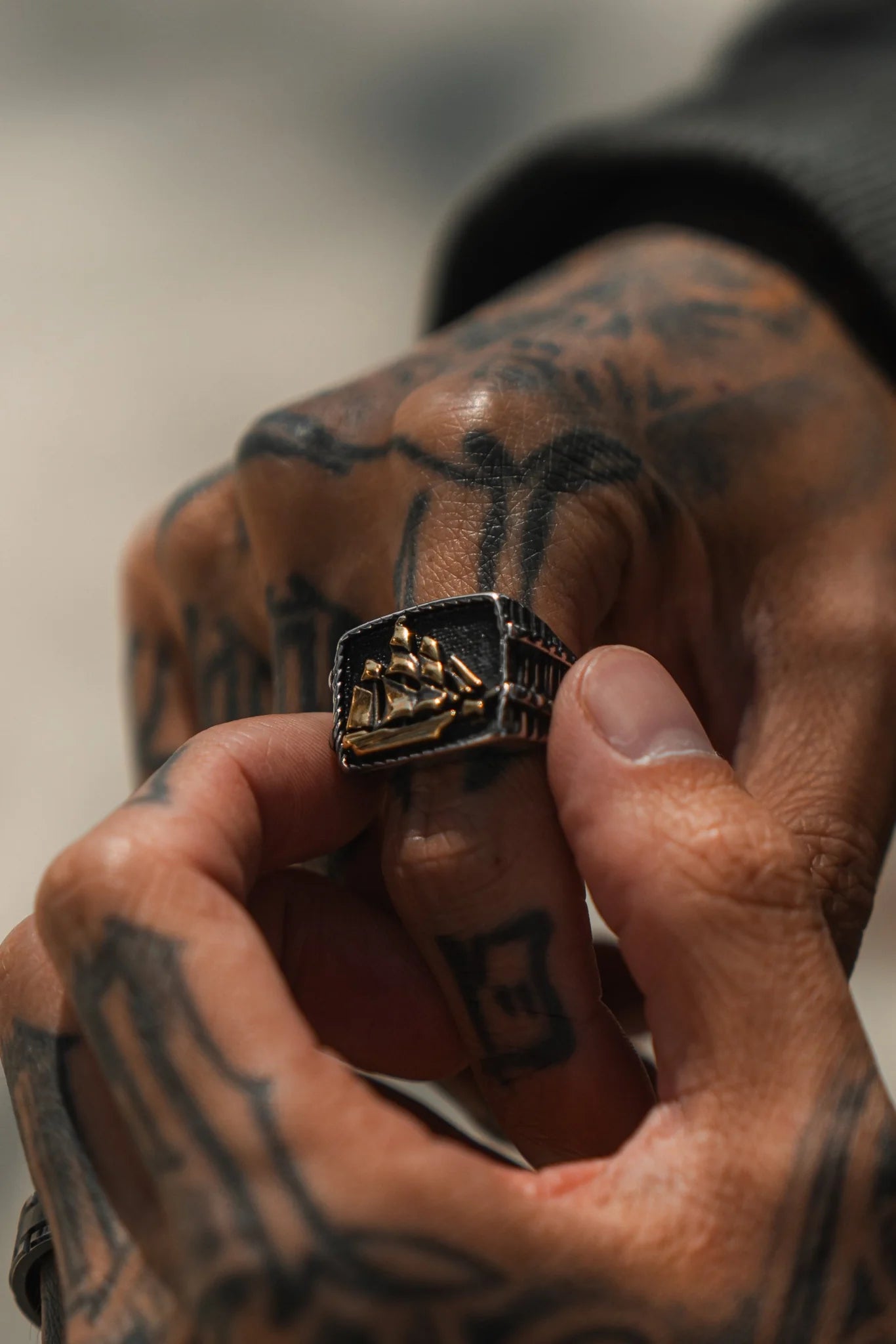 A tattooed hand holds the Queen Annes Revenge Ring by Relentless Betrayal, featuring a dark band and raised golden ship design. This unique piece evokes pirate lore and Blackbeards famed vessel, set against a blurred background that emphasizes its intricate details.