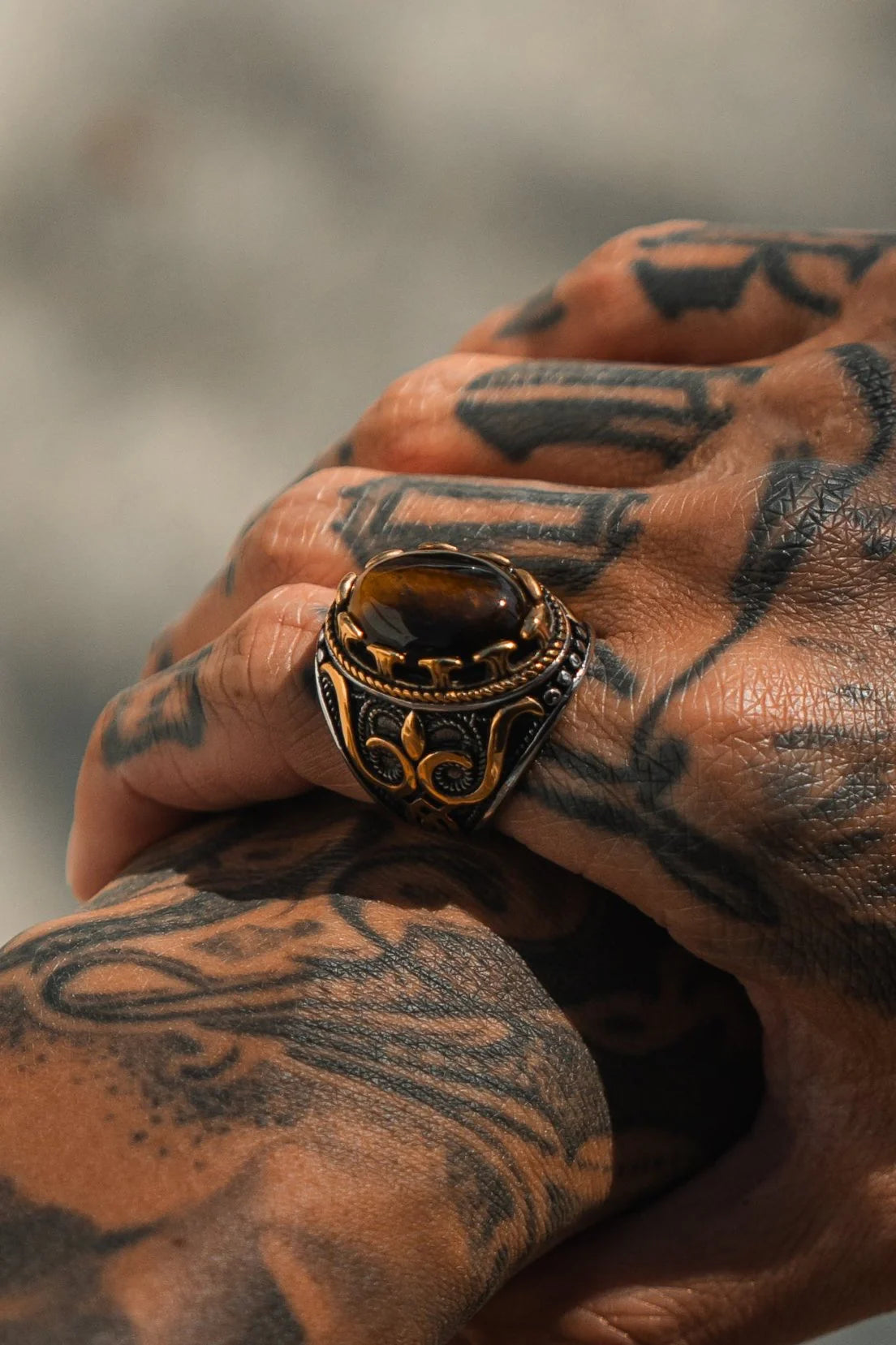 A hand with intricate tattoos displays the Royal Revenge Ring by Relentless Betrayal, an ornate heirloom with a glossy brown gemstone and decorative patterns. The lighting accentuates the rings and tattoos textures and details beautifully.