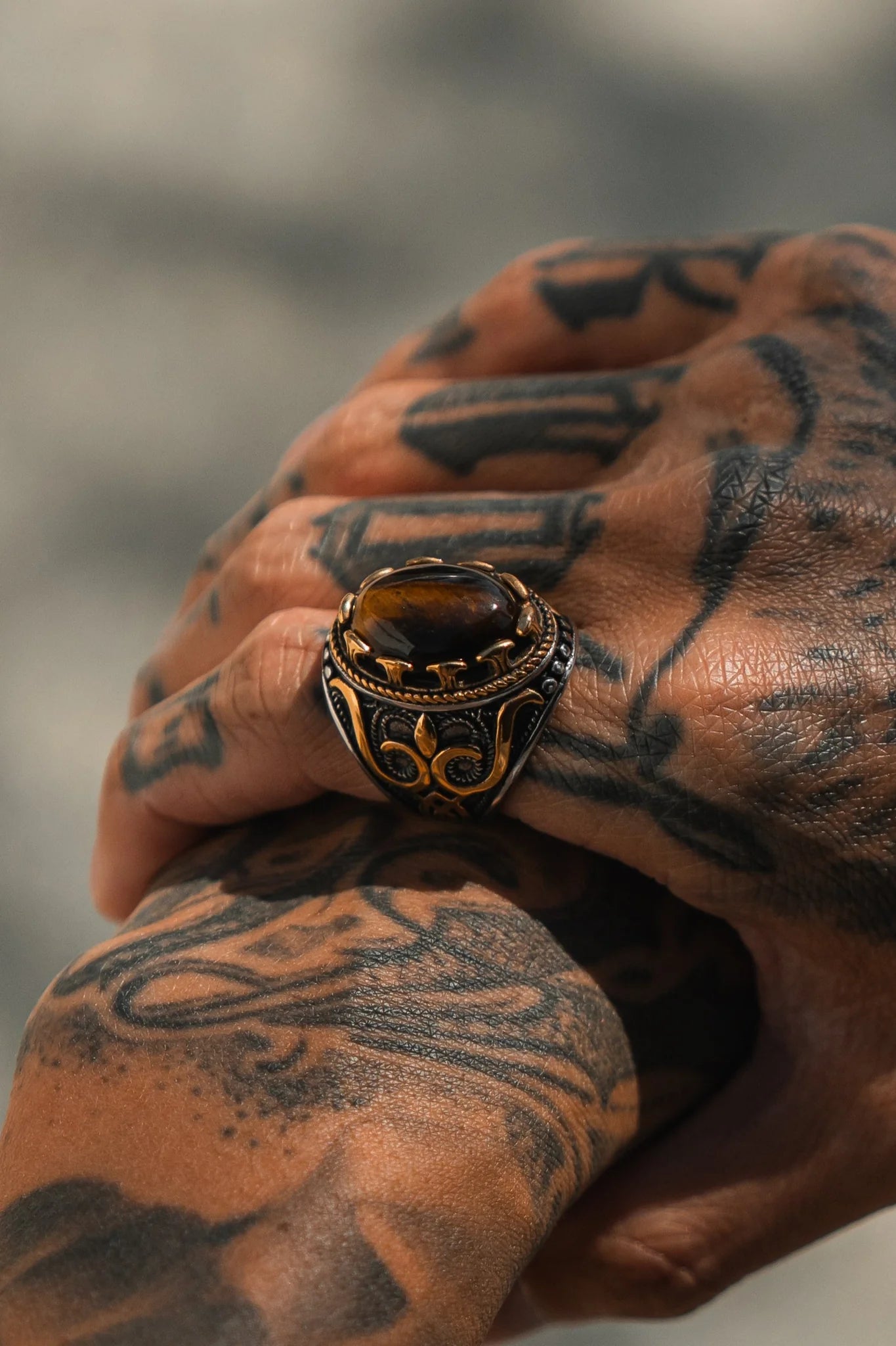 A tattooed hand displays the Royal Revenge Ring by Relentless Betrayal, featuring a large dark stone and intricate patterns with gold accents. The handcrafted heirlooms detailed design stands out against the blurred background.