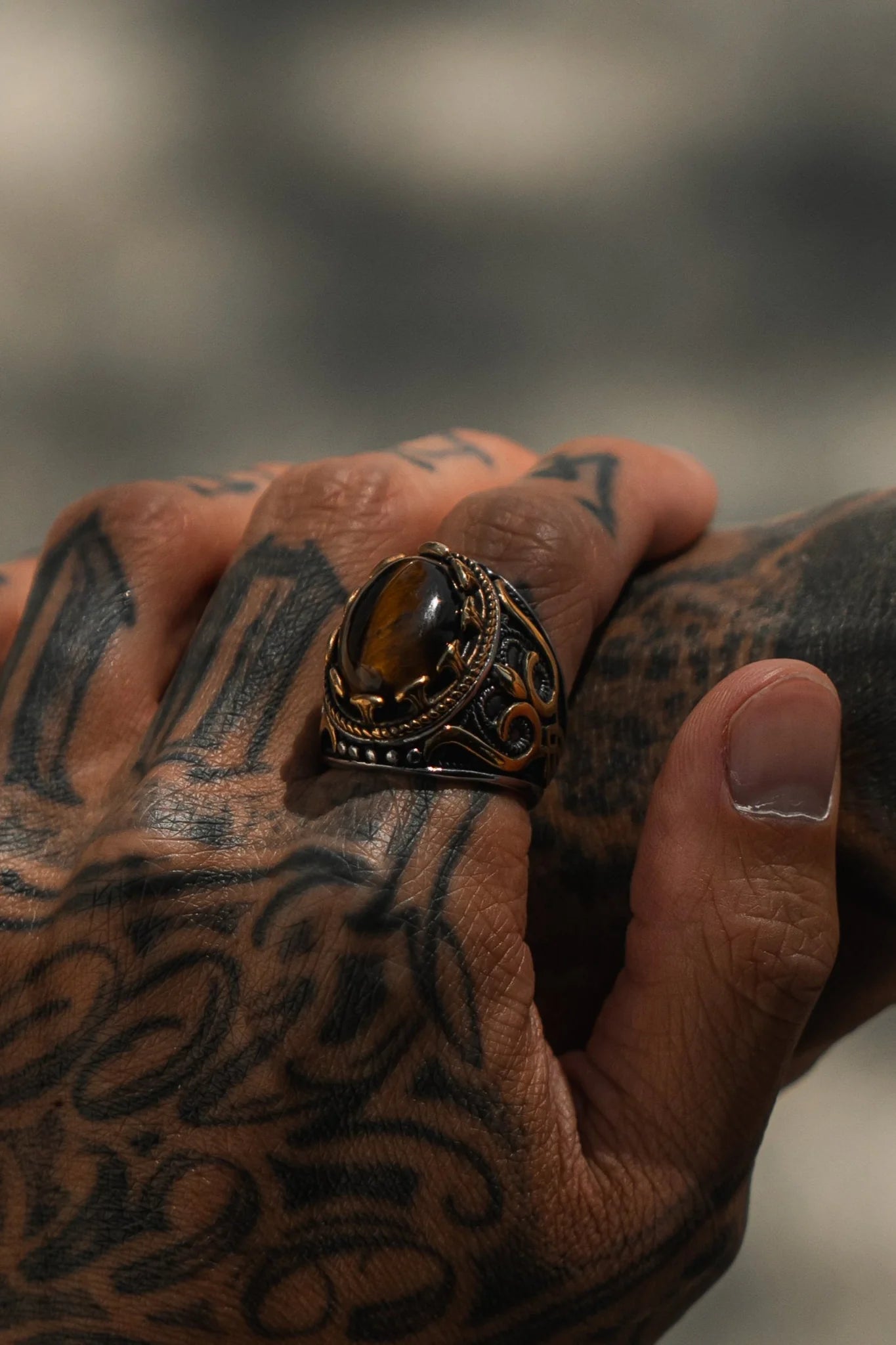 A hand with intricate tattoos holds a dark object adorned with the Royal Revenge Ring by Relentless Betrayal. This heirloom piece flaunts a prominent brown gemstone in an ornate antique-style metal band against a blurred background, enhancing the hands elegant mystique.
