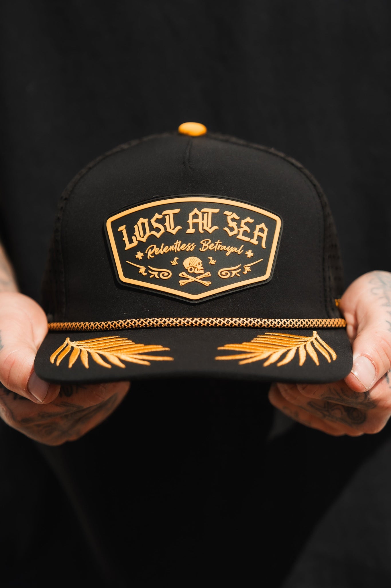 A person holds a Lost At Sea PVC Snapback by Relentless Betrayal, featuring gold lettering and a brim adorned with gold leaf embroidery. This water-resistant black cap is the perfect mix of durability and style for a pirate lifestyle against a dark, neutral backdrop.