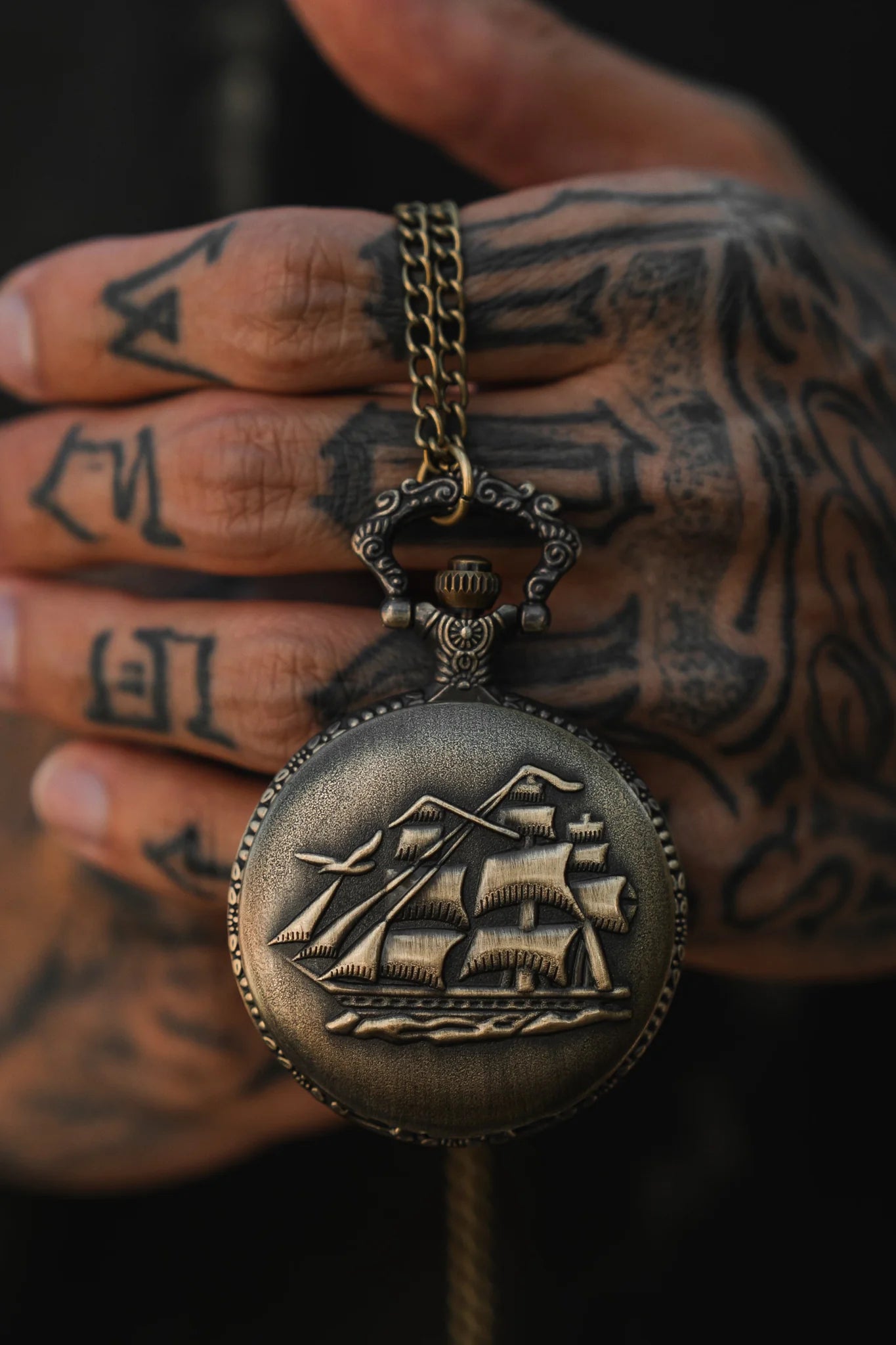 A tattooed hand holds The Legacy Pocket Watch by Relentless Betrayal, with a pirate ship design evoking adventure. Its stainless steel chain drapes elegantly, creating a dark, mysterious tone.