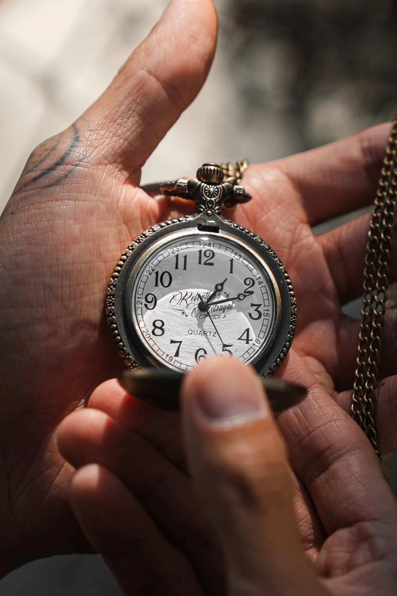 A person holds The Legacy Pocket Watch by Relentless Betrayal, featuring an elegantly etched pirate ship and a chain. The time shows 1:51. A tattoo adorns the wrist, while a softly blurred background keeps focus on the watch and hand.