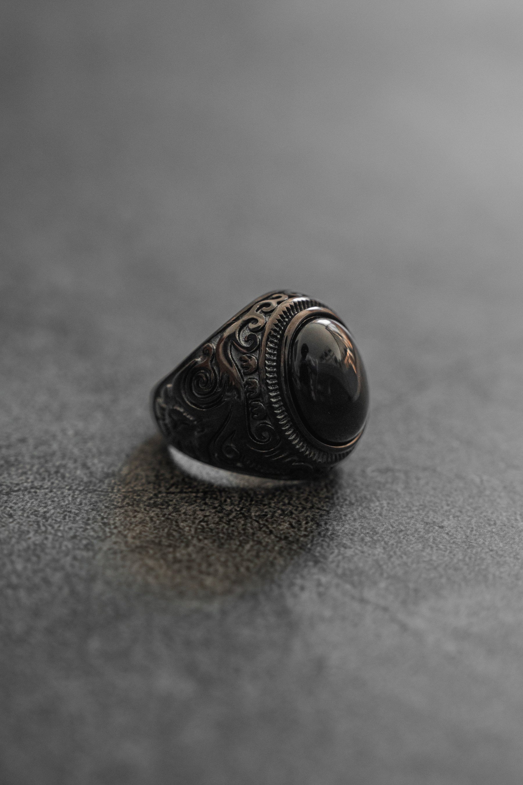 Relentless Betrayals Black Pearl Ring, featuring intricate swirling patterns and a large oval black stone, elegantly rests on a textured gray surface with a softly blurred background that highlights the stainless steel jewelrys ornate details.