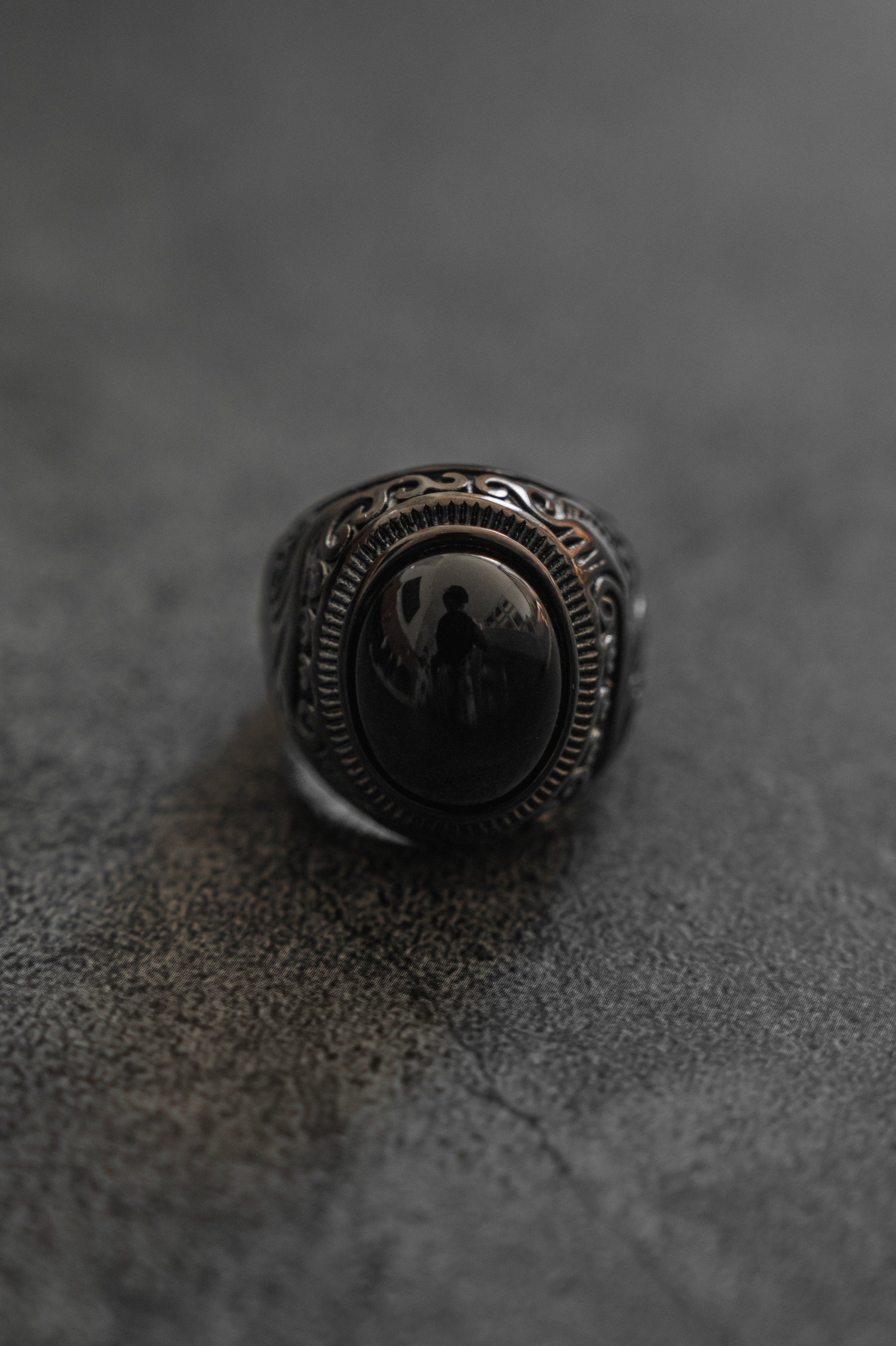 A detailed view of Relentless Betrayals The Black Pearl Ring displays a dark, intricate design featuring an oval black stone in a complex silver band, inspired by French Polynesia pearls, on a textured dark gray surface. The stone’s reflection captures a faint silhouette.