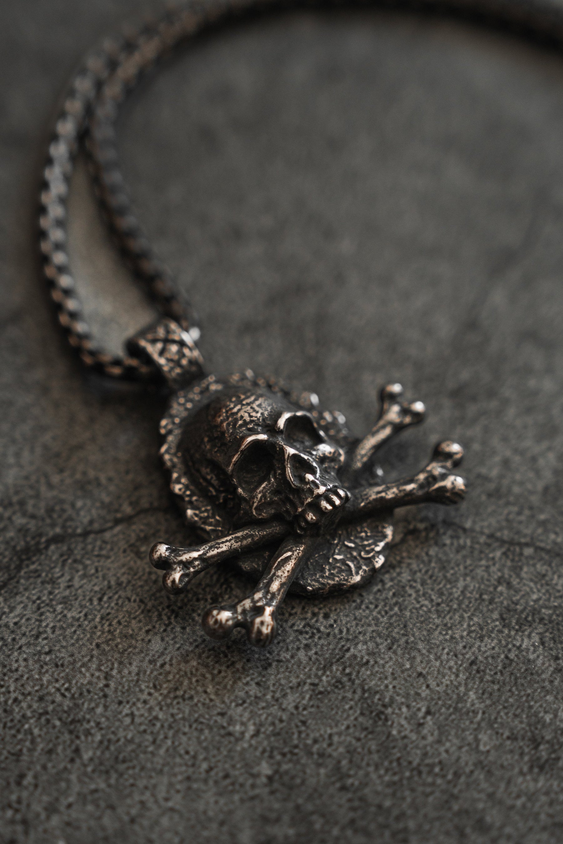 The Tell No Tales Necklace by Relentless Betrayal rests on a textured dark surface, featuring a skull and crossbones pendant. Its sturdy stainless-steel chain complements the intricate design.
