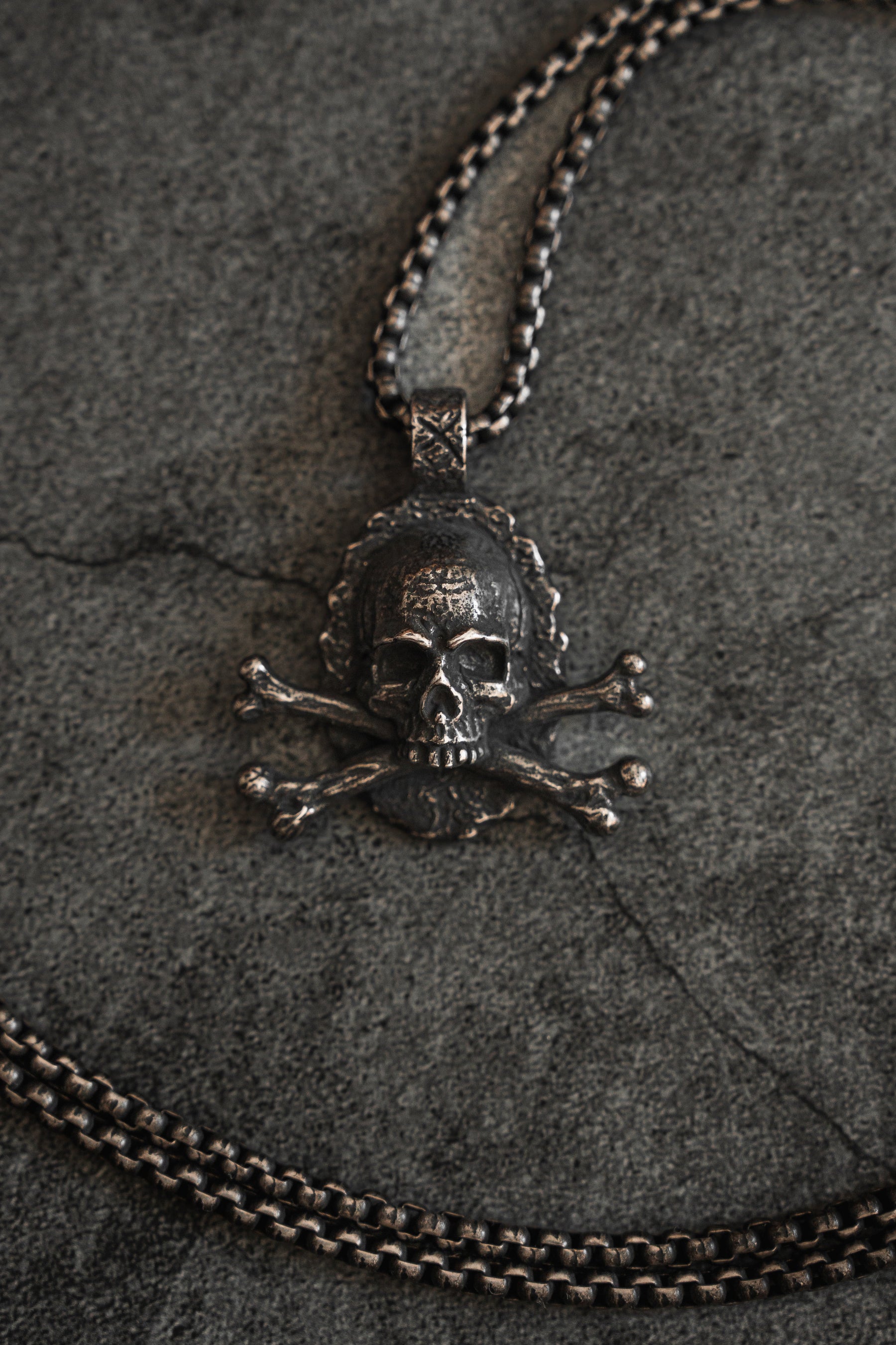 The Tell No Tales Necklace by Relentless Betrayal sits on a dark, textured surface, showcasing a handcrafted skull and crossbones pendant. Its intricate metallic details and hand-forged stainless steel design create an edgy yet elegant look.