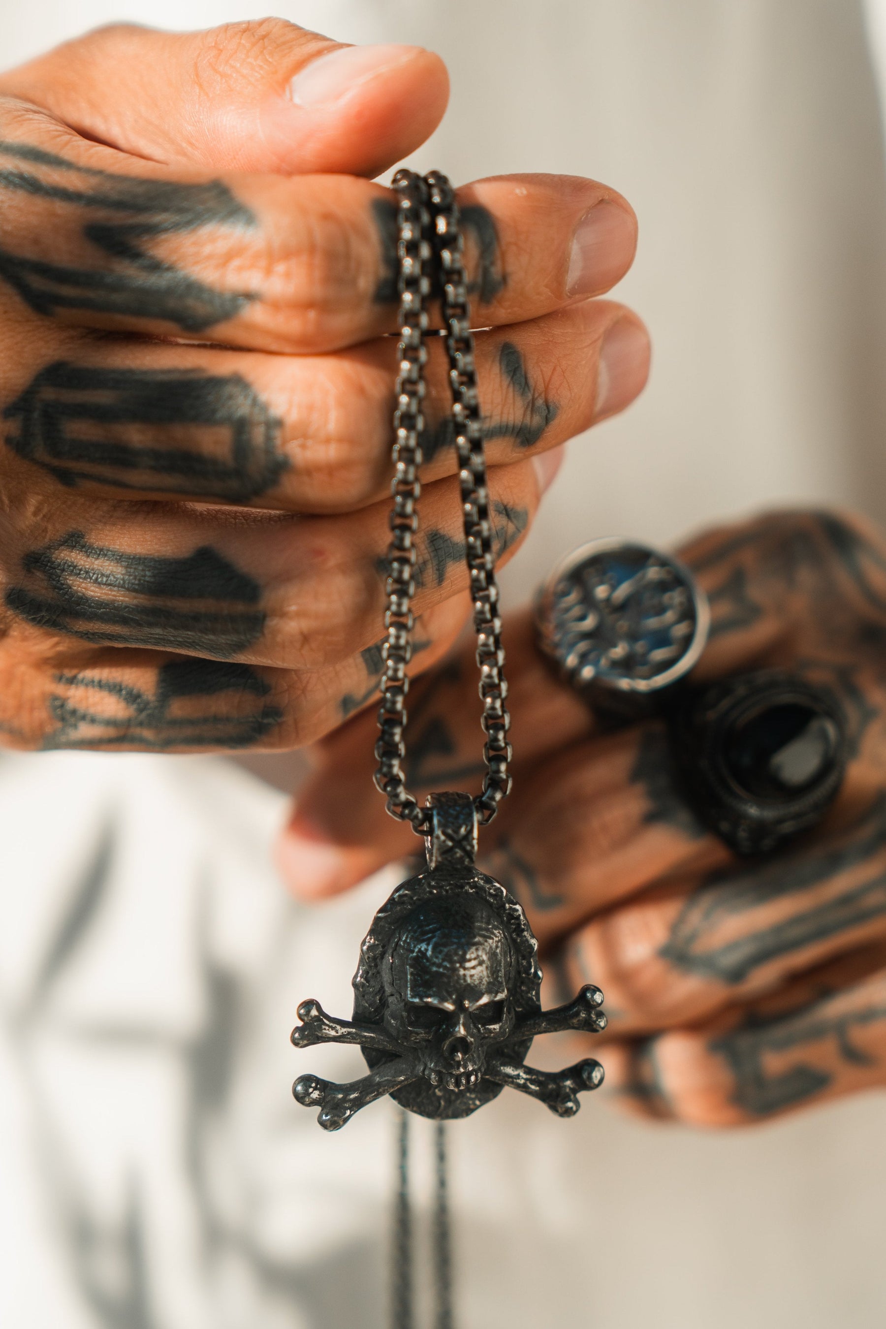 A close-up of tattooed hands with large rings holds the Tell No Tales Necklace by Relentless Betrayal, featuring a skull and crossbones pendant on a hand-forged stainless steel chain. The dark tattoos cover most of the fingers and hands.