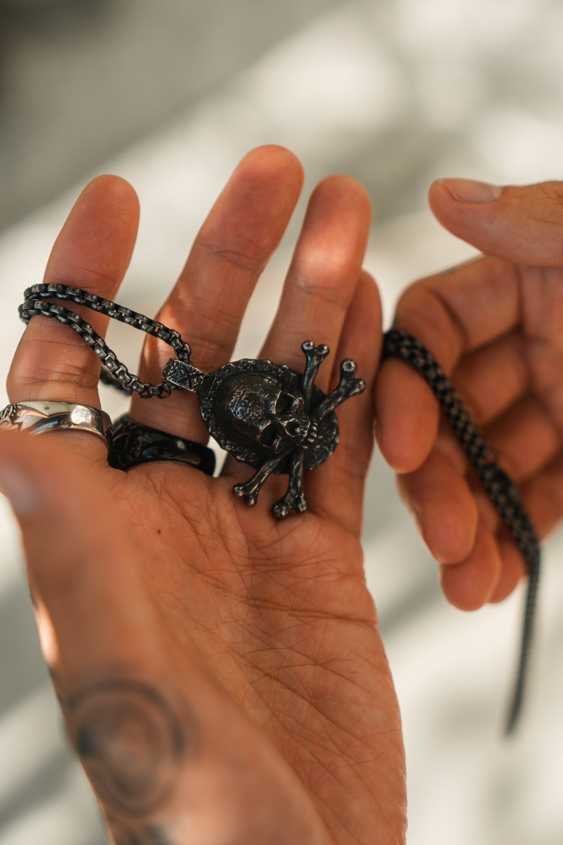 A hand holds the Tell No Tales Necklace by Relentless Betrayal, showcasing a skull-shaped pendant on a stainless-steel chain against a light background. Fingers adorned with rings and a tattoo cast lively shadows across the surface.