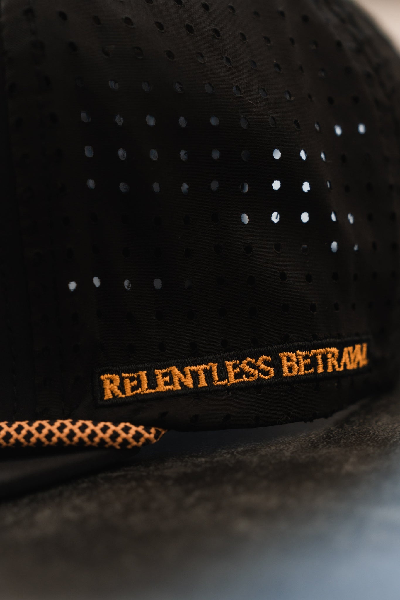 Close-up of the Lost At Sea PVC Snapback by Relentless Betrayal, a black cap with small holes, featuring an orange embroidered patch reading RELENTLESS BETRAYAL and a textured orange detail near the brim. Perfect for the pirate lifestyle, this snapback is water-resistant.