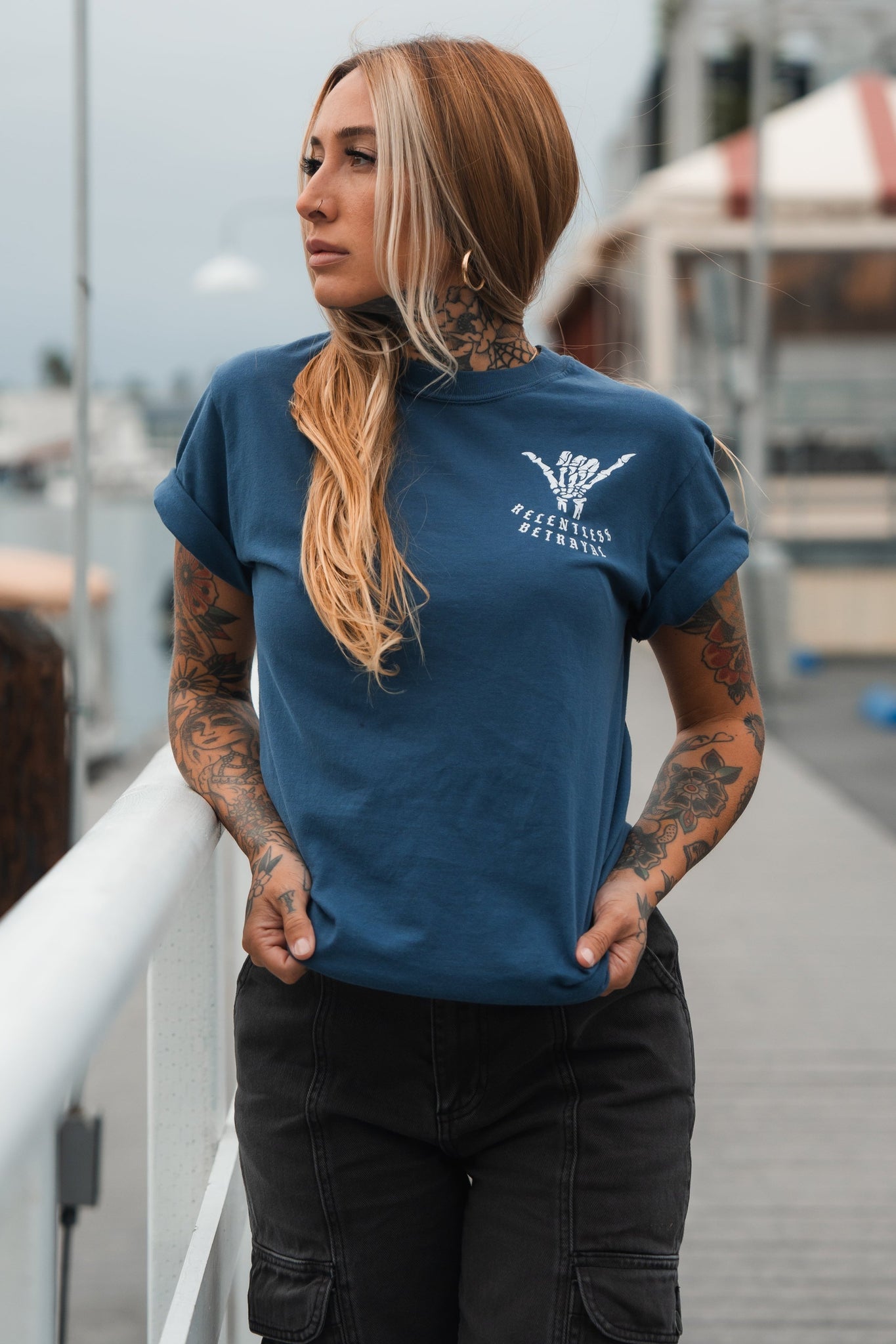 A tattooed woman with long hair, wearing a blue Sink or Swim Premium Slate T-Shirt by Relentless Betrayal, stands on a pier with hands in pockets. She gazes sideways resiliently against an overcast sky and distant buildings.