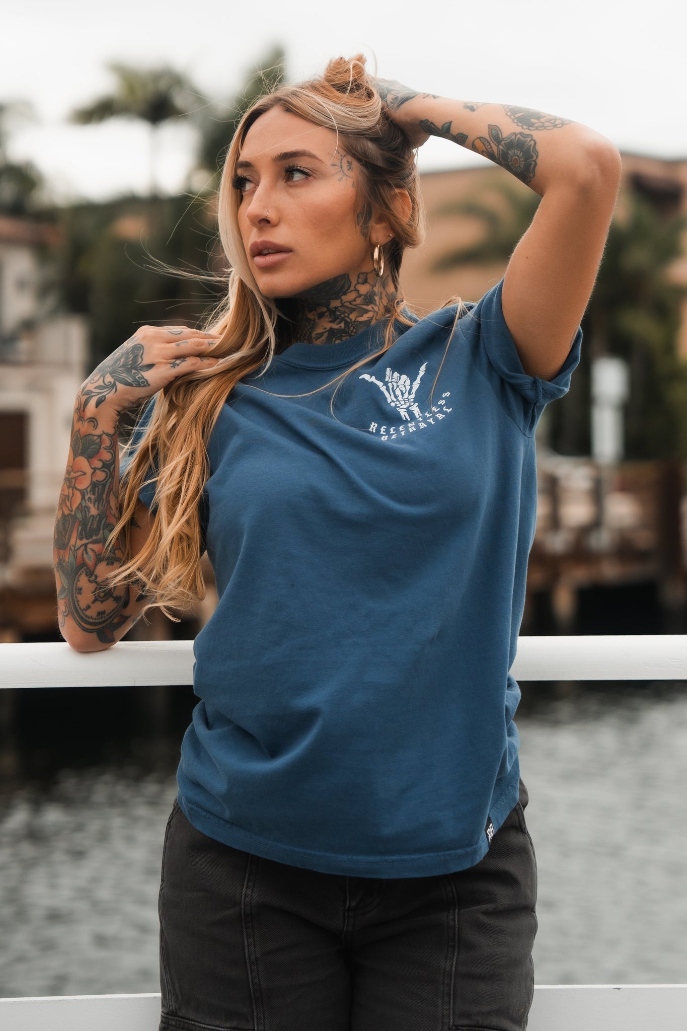 A person stands resiliently on a bridge, touching their head with one hand, wearing a Relentless Betrayal Sink or Swim Premium Slate T-Shirt and dark pants. Their neck and arm tattoos exude inner strength, while the blurred water, trees, and buildings create a serene backdrop.