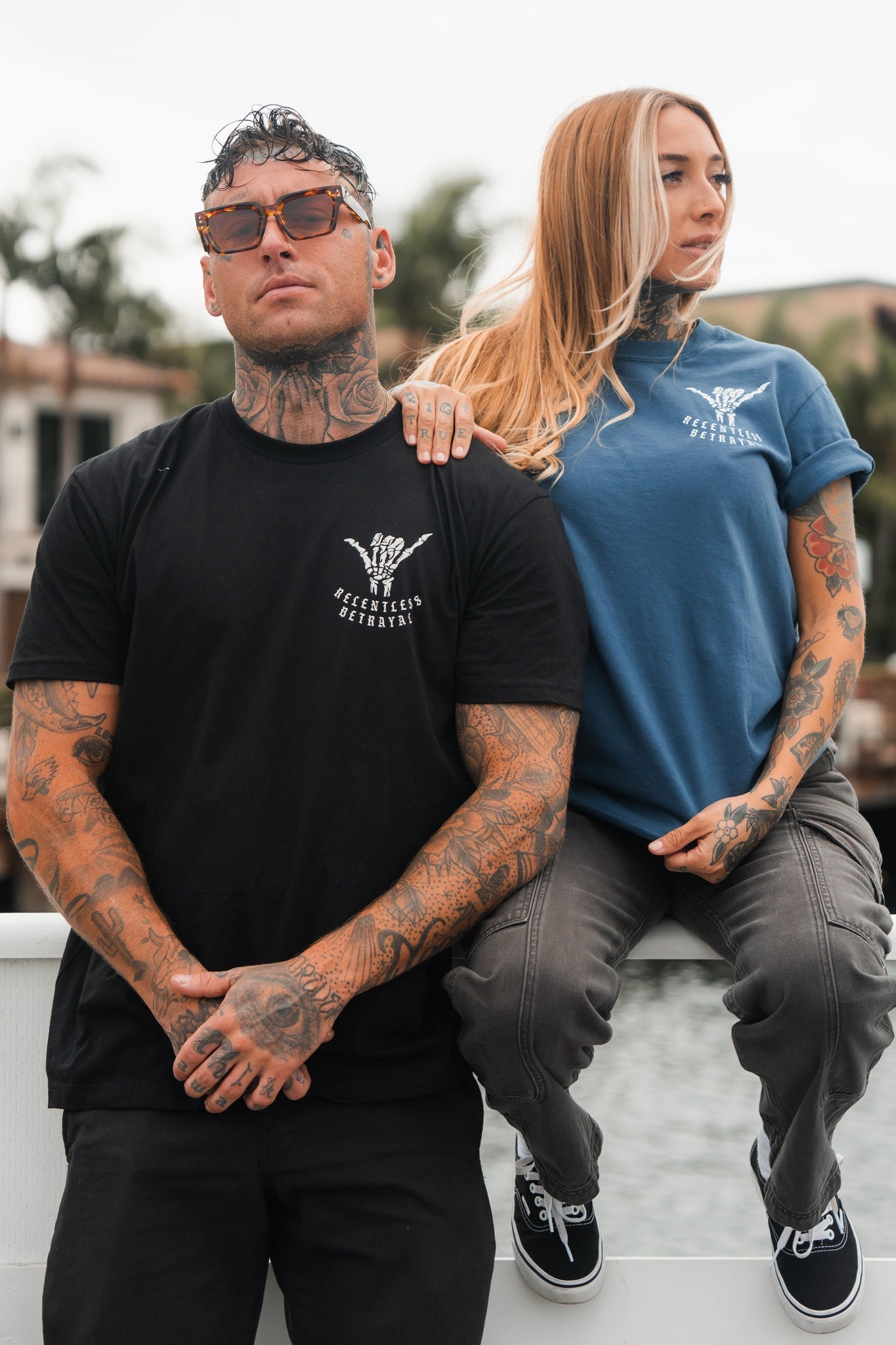A person flaunting tattoos and wearing a Relentless Betrayals Sink or Swim Tee - Black, stands confidently in sunglasses. Next to them, an individual with long hair and tattoos sits on a railing in a blue shirt. Both exude style and embody perseverance through life’s challenges.