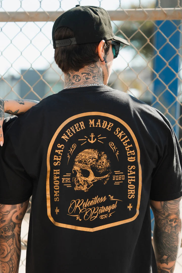 A person with tattoos wears the Skilled Sailor Premium Gold Foil T-Shirt by Relentless Betrayal, made from premium jersey cotton, featuring skull and nautical imagery and the text Smooth seas never made skilled sailors. A chain-link fence is in the background.