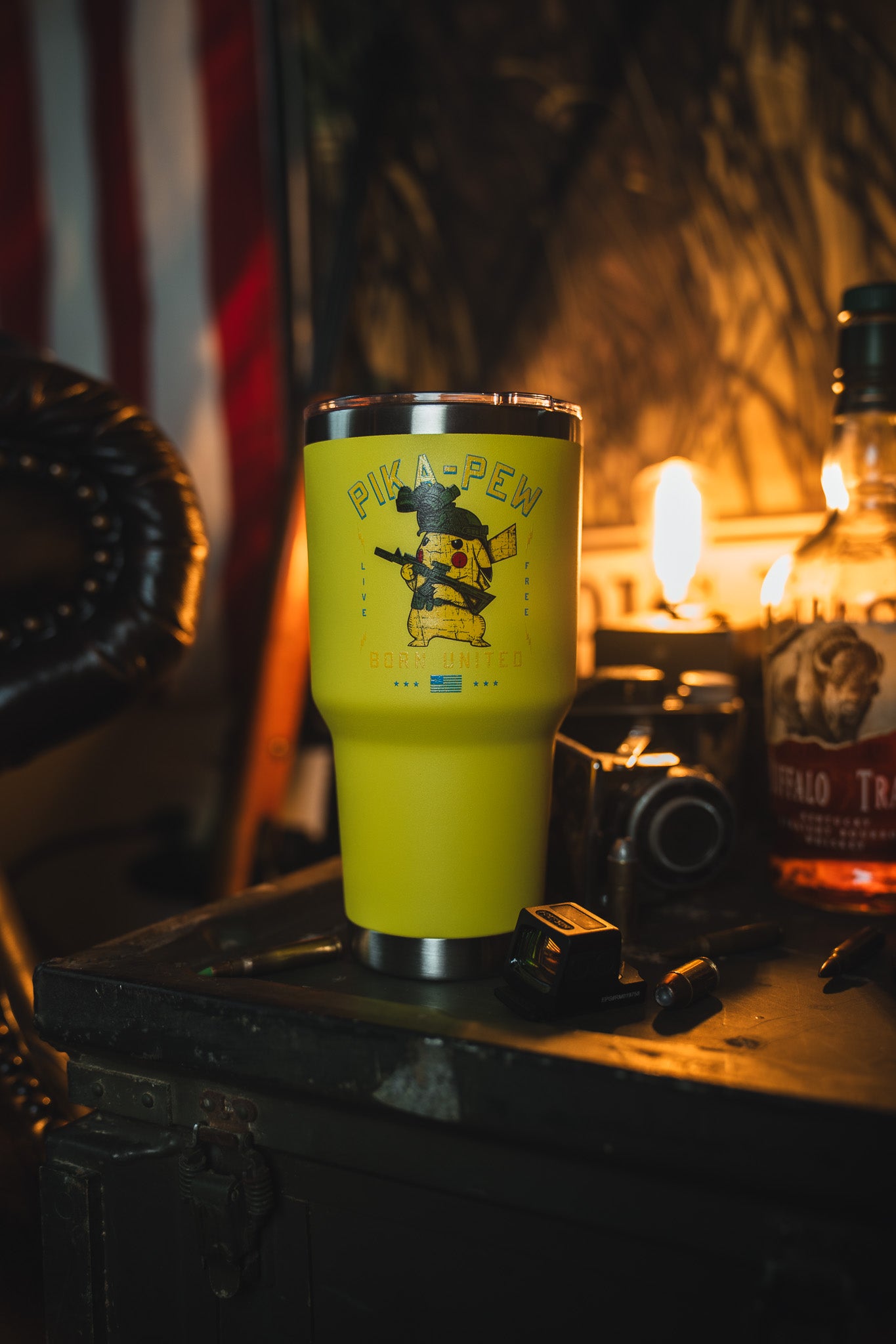 The 30oz Pika-Pew Tumbler by Born United Private Label—bright yellow with a cartoon mouse holding guns—rests on a table in a warmly lit, rustic room alongside a camera, whiskey bottle, and scattered items.
