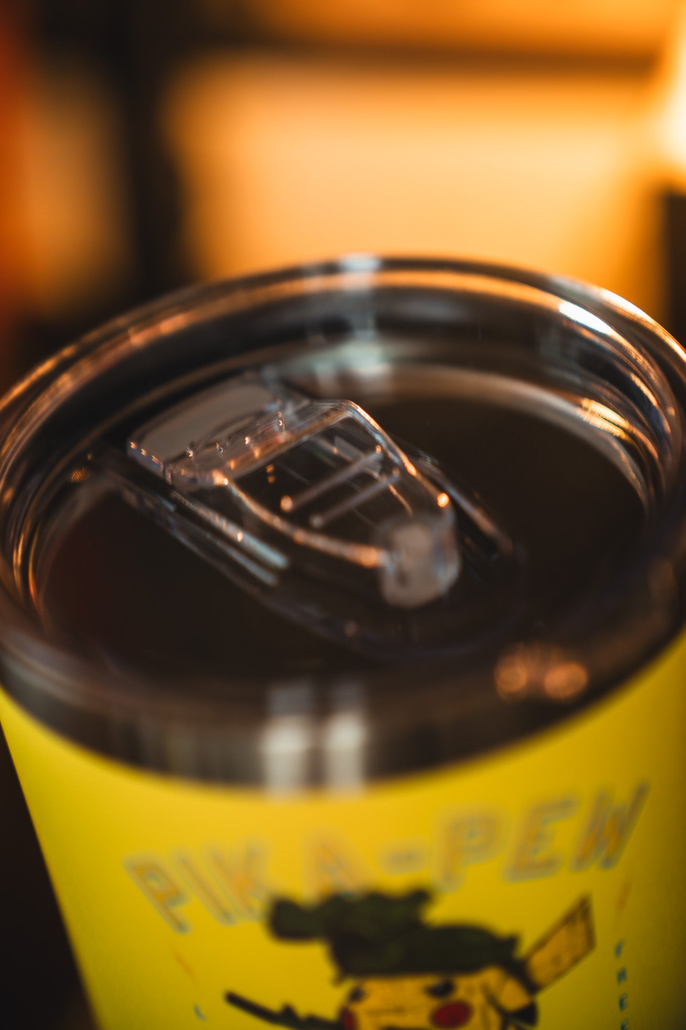 A close-up of the Born United Private Label Pika-Pew Tumbler - 30oz with a clear plastic lid, yellow sleeve featuring a Pikachu illustration, and warm lighting reflecting on its surface.