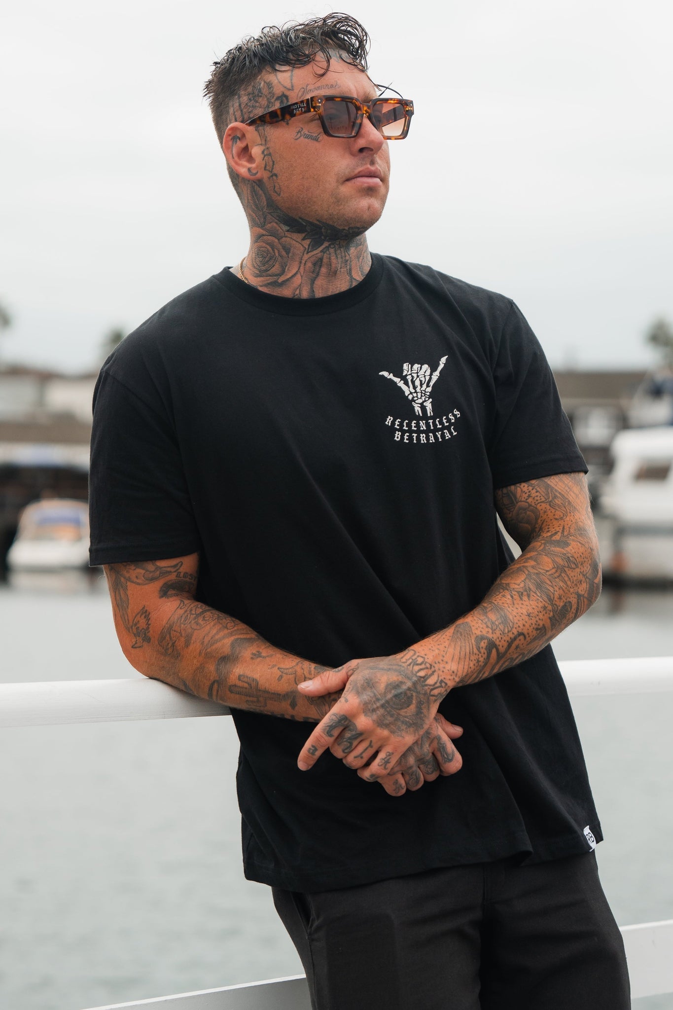 A tattooed man exuding resilience stands by a railing in his Relentless Betrayal Sink or Swim Tee - Black and sunglasses. The marinas blurred boats symbolize the challenges he navigates with perseverance.