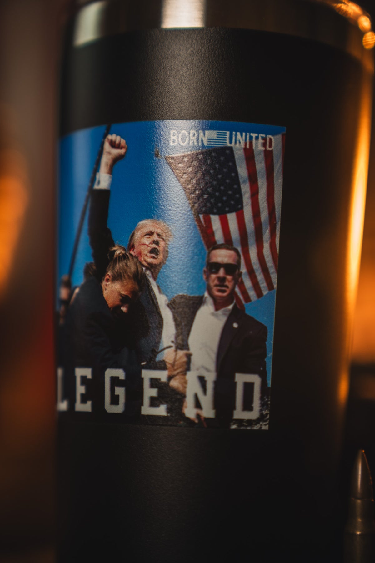 The Forever Legend Tumbler - 20oz by Born United Private Label features a sticker with a suited man raising his fist, a man in sunglasses, and a bowed woman before an American flag. The sticker says BORN UNITED and LEGEND.