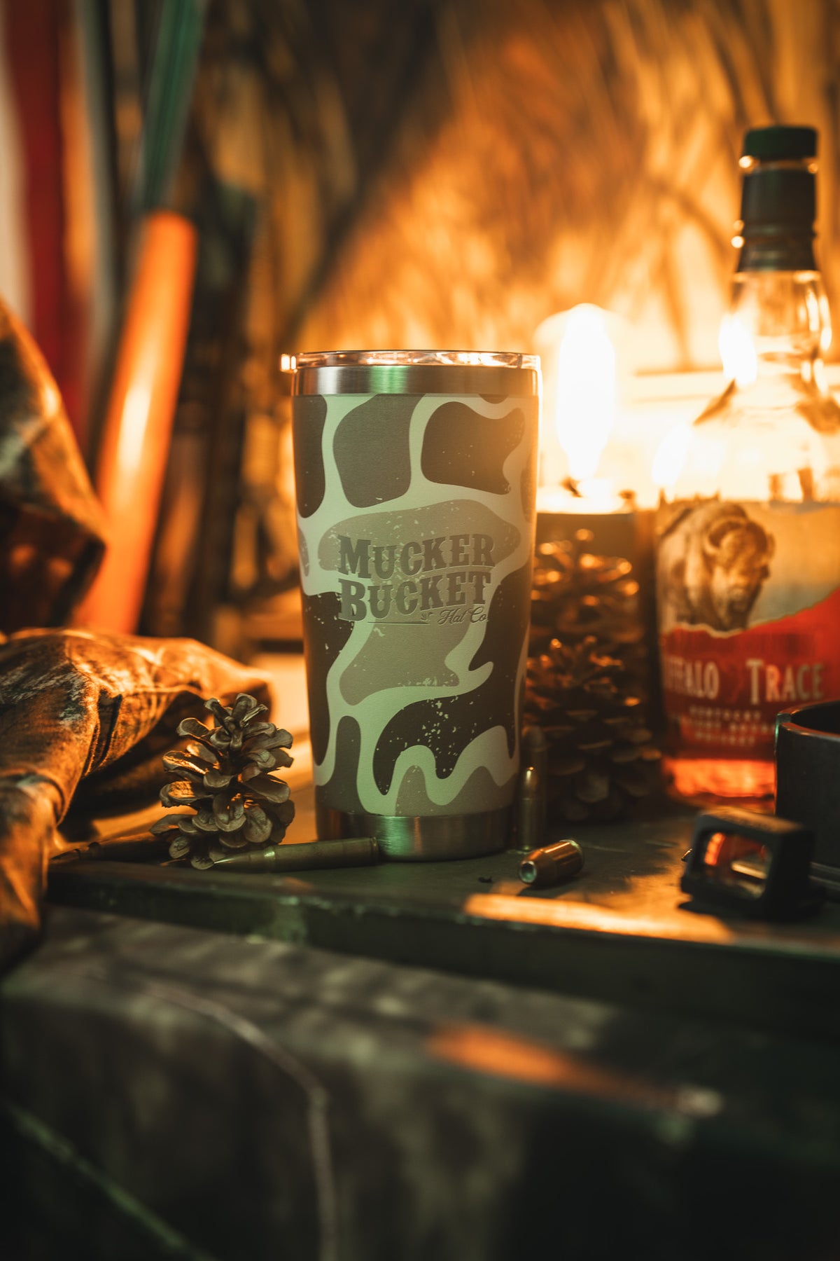 The Mucker Bucket Duck Camo Tumbler - 20oz sits beside a pine cone, bullet casing, and Buffalo Trace whiskey under warm light—a laser-engraved stainless steel tumbler ideal for any rustic setting.