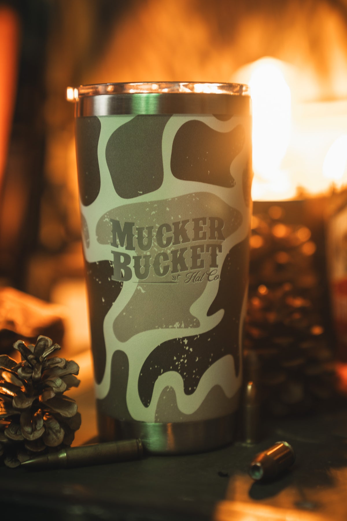 The Mucker Bucket Duck Camo Tumbler - 20oz rests beside pinecones and a bullet casing under warm, blurry light. Its laser-engraved stainless steel design combines outdoor charm with rugged style.