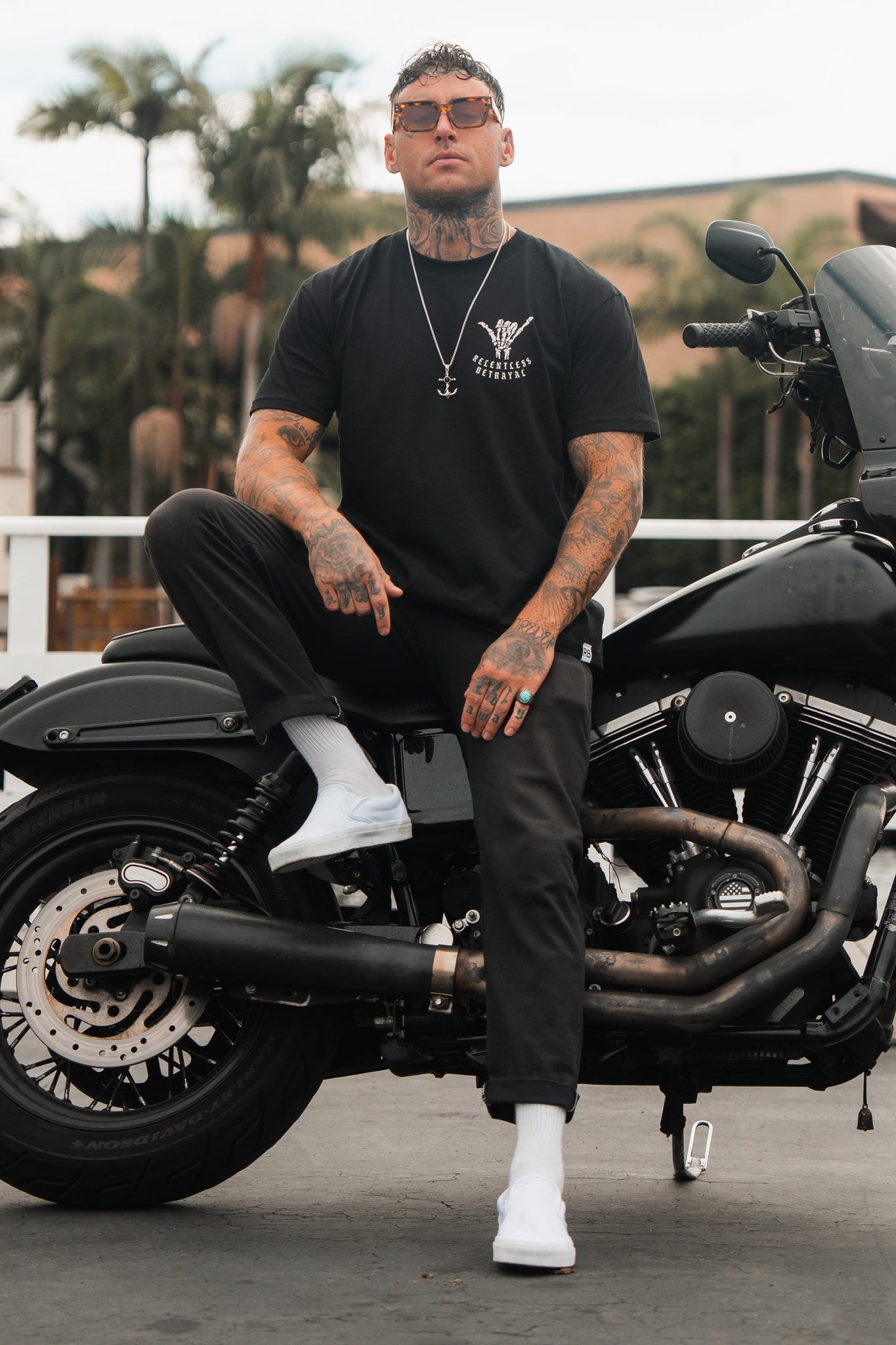 Sporting a Sink or Swim Tee by Relentless Betrayal, a tattooed man embodies resilience on his black motorcycle. He wears sunglasses, dark pants, white socks, and shoes against a backdrop of palm trees and buildings, reflecting his steadfast perseverance through challenges.
