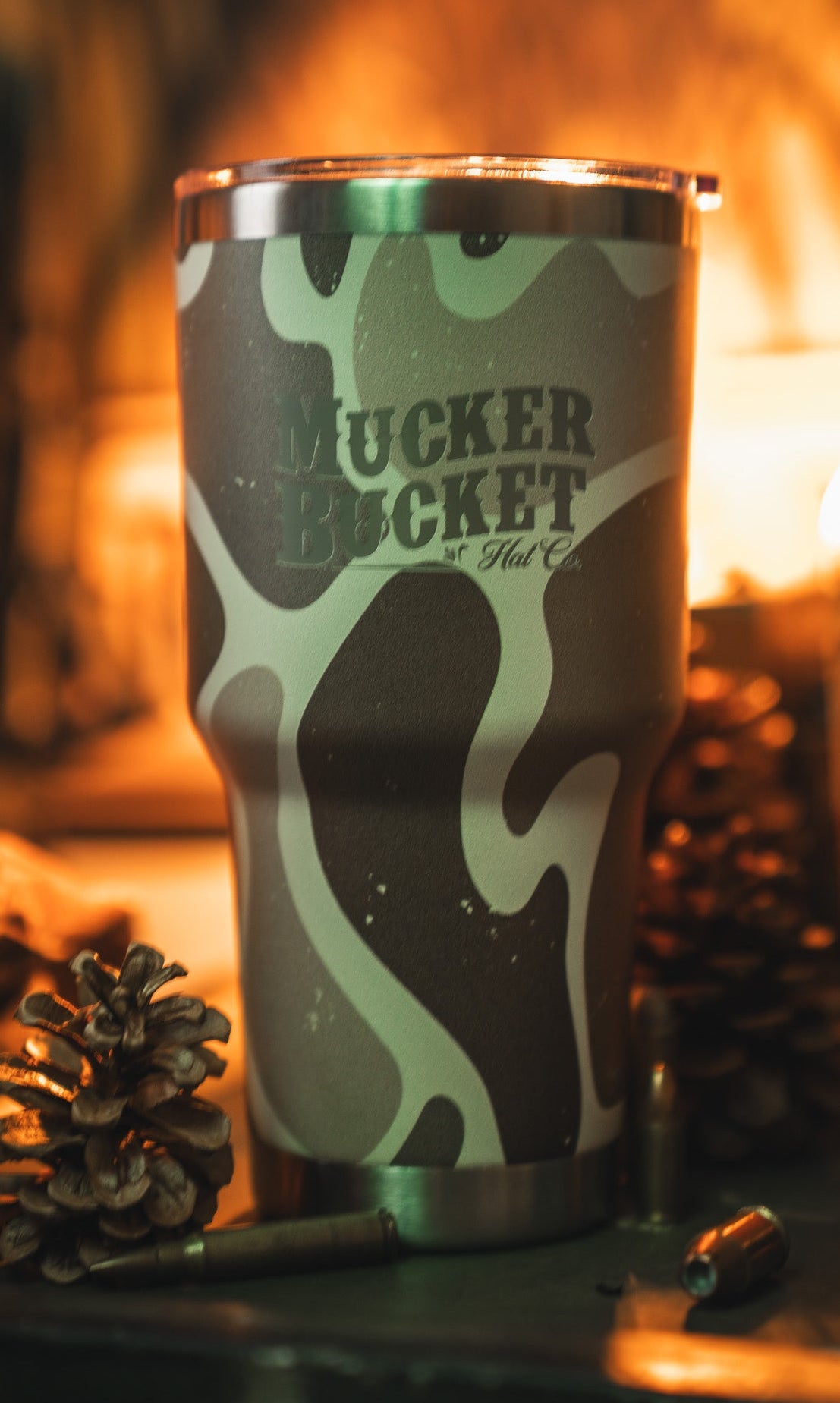The Duck Camo Tumbler - 30oz by Mucker Bucket features a green and tan camouflage design and custom laser engraving, displayed with pinecones, a bullet, and warm ambient lighting.