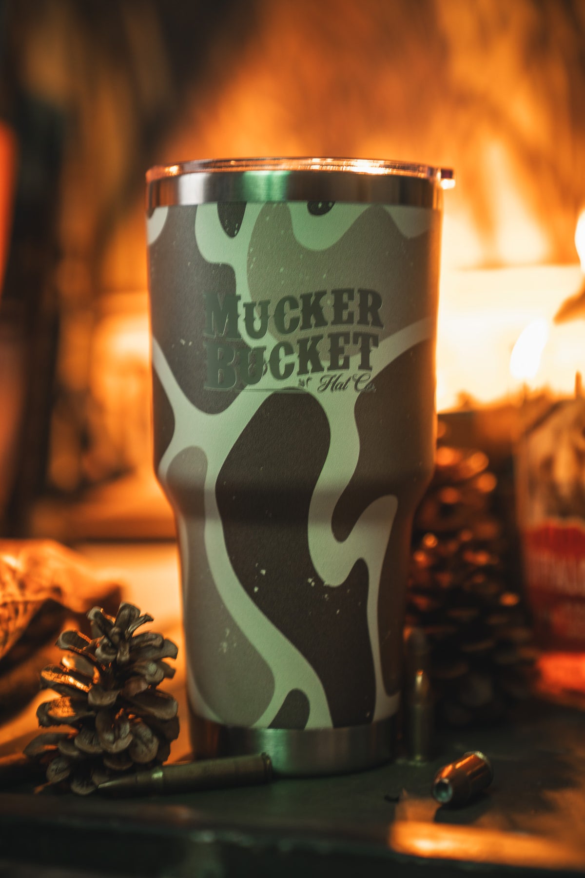 The Duck Camo Tumbler - 30oz by Mucker Bucket features a green and tan camouflage design and custom laser engraving, displayed with pinecones, a bullet, and warm ambient lighting.