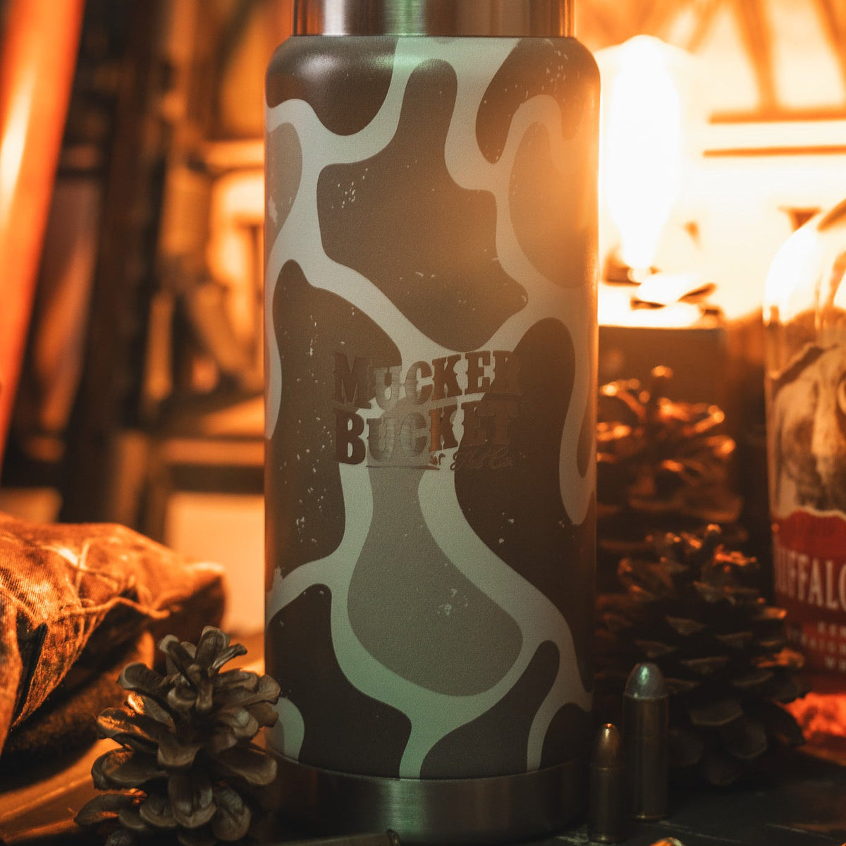 A close-up of the 26oz Duck Jug - Duck Camo Water Bottle by Mucker Bucket sits on a table with pinecones, a metal bullet, and a bottle in warm, soft lighting.