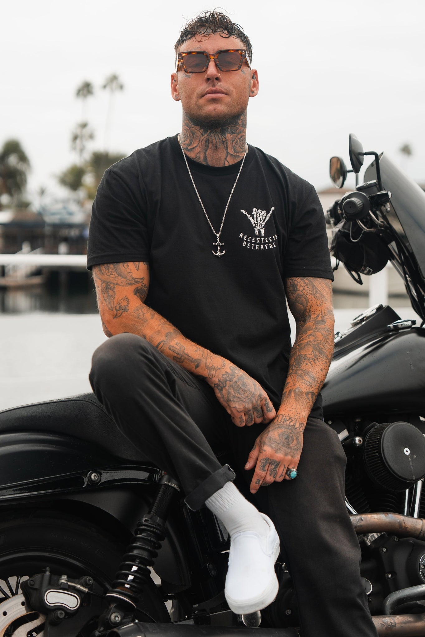 A tattooed man, embodying resilience, wearing Relentless Betrayals Sink or Swim Tee - Black, sunglasses, and white shoes, sits on a black motorcycle near a waterfront with palm trees and boats.