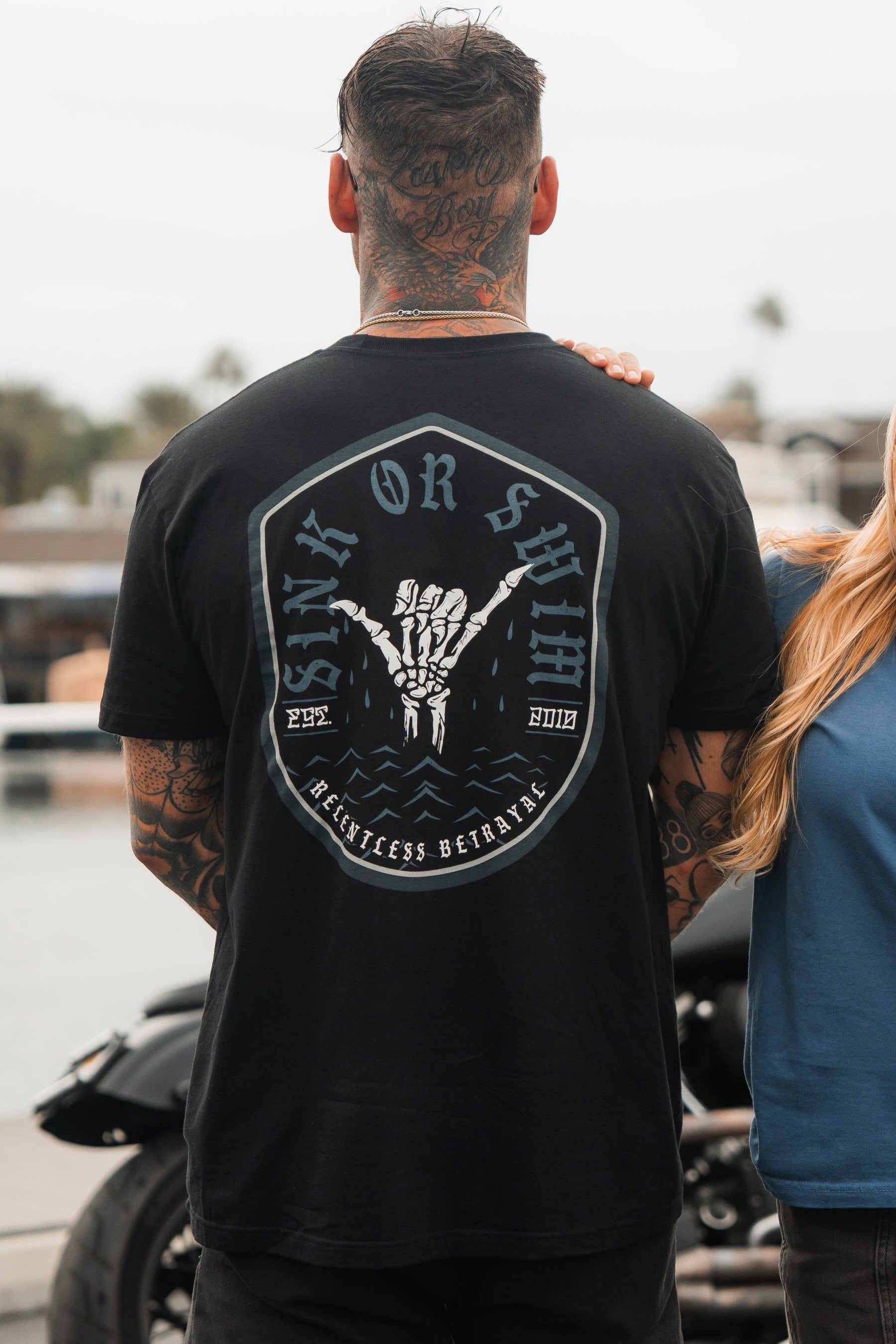 A person with tattoos faces away from the camera in a black Sink or Swim Tee by Relentless Betrayal, featuring a hand graphic and phrase symbolizing resilience. Another person partially appears beside them.