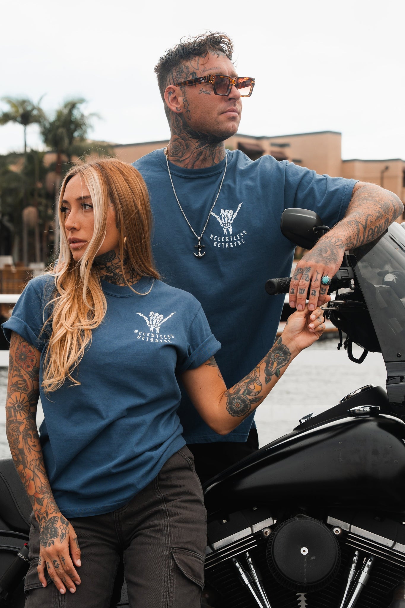 Two tattooed individuals stand by a motorcycle in blue Sink or Swim T-shirts by Relentless Betrayal, featuring white designs with text. The man wears sunglasses and leans on the bike, while the woman rests her hand on his arm. Palm trees and buildings provide the backdrop.