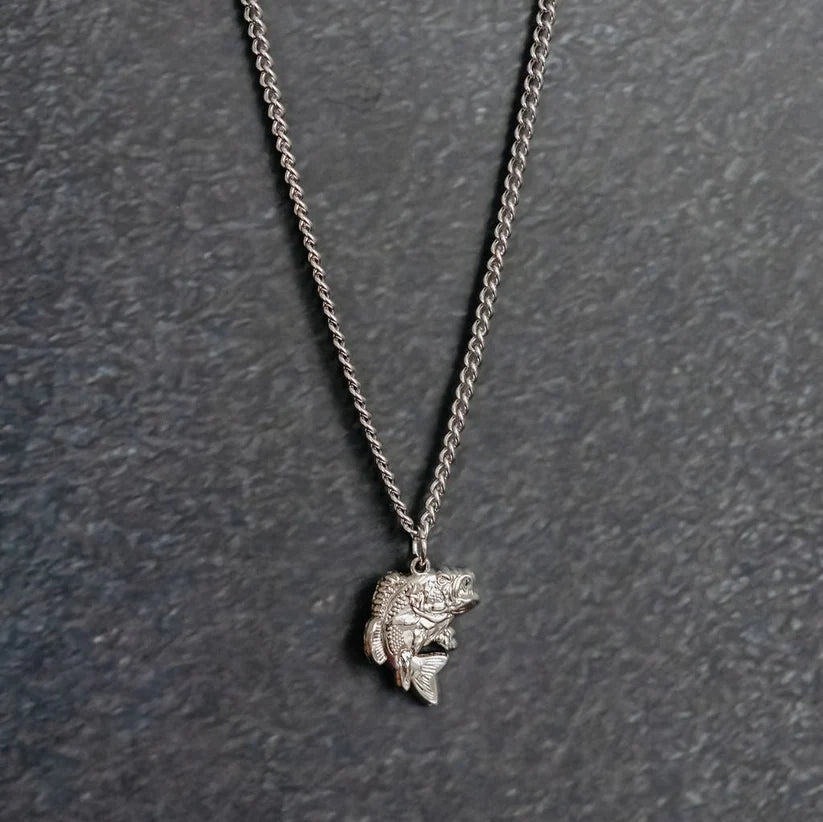 The Big Bass America Necklace by Mucker Bucket is a silver piece featuring a fish-shaped pendant, showcased against a textured dark gray backdrop.