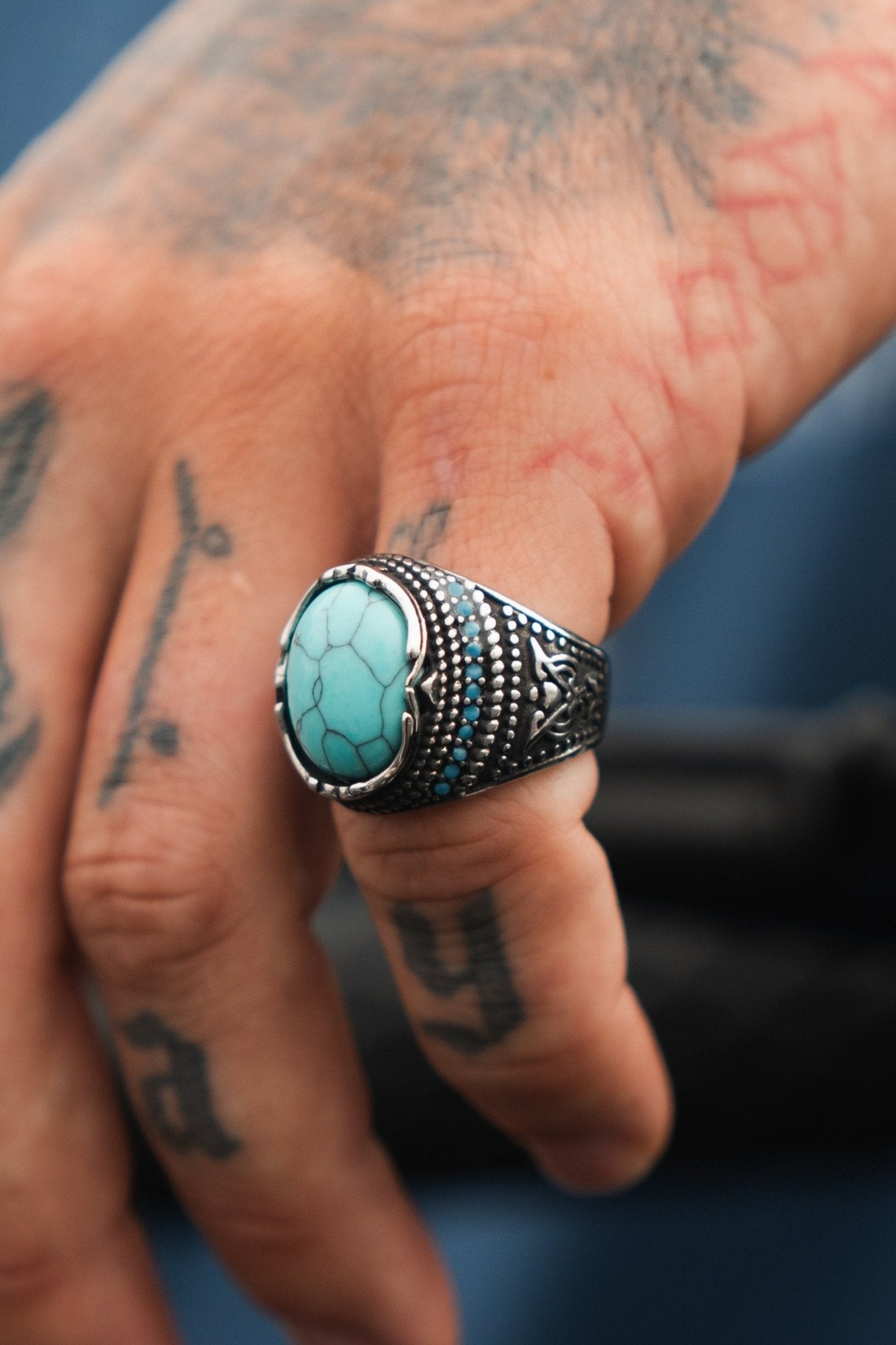 A tattooed hand displays the ornate Eye of the Storm Ring by Relentless Betrayal, featuring a turquoise stone and intricate metalwork. The blurred background emphasizes this exquisite handcrafted jewelry piece.
