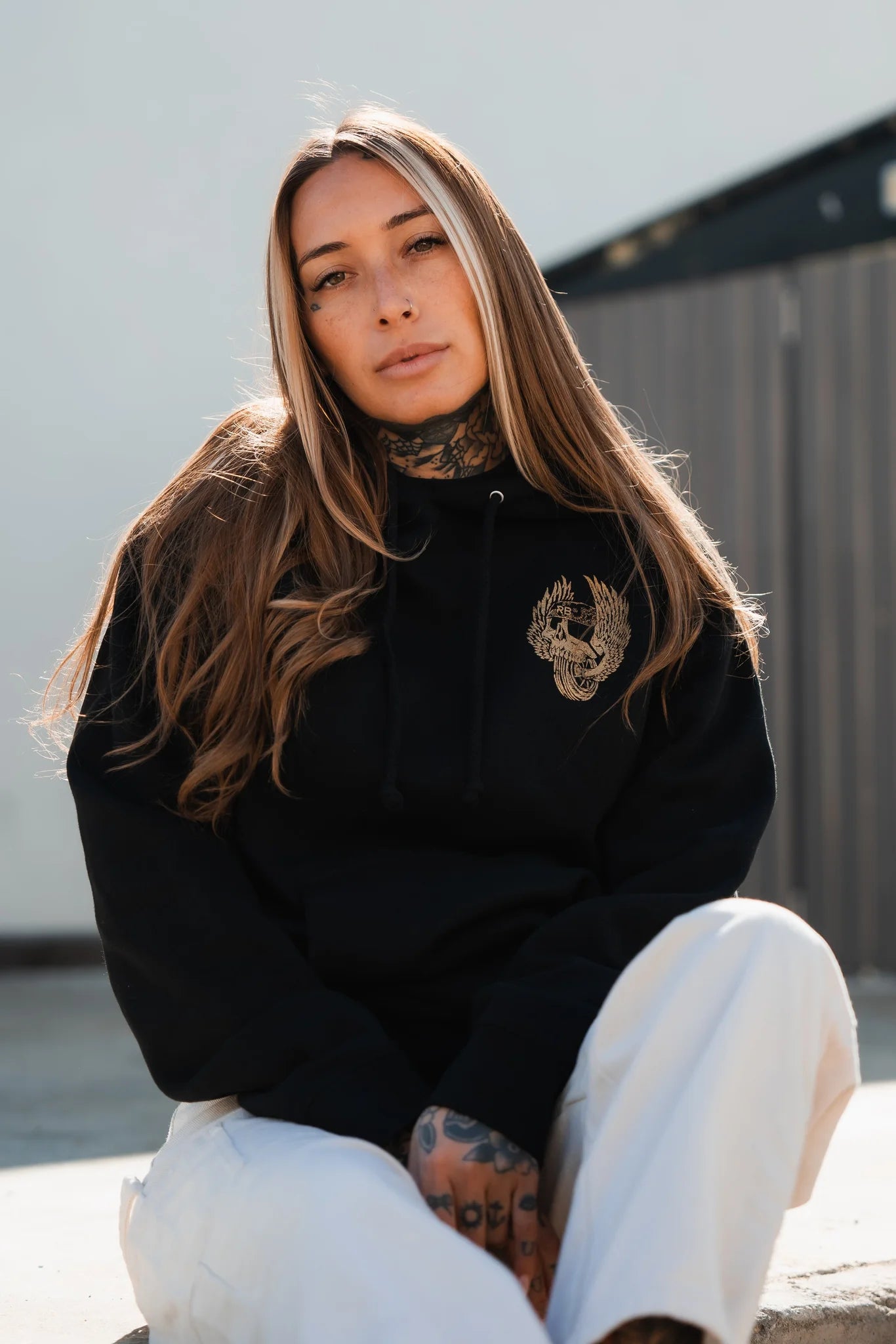 Sitting outdoors, a long-haired, tattooed person embodies youth culture in a black Relentless Betrayal All Gas No Brakes Premium Hoodie and white pants, looking directly at the camera with a neutral expression.