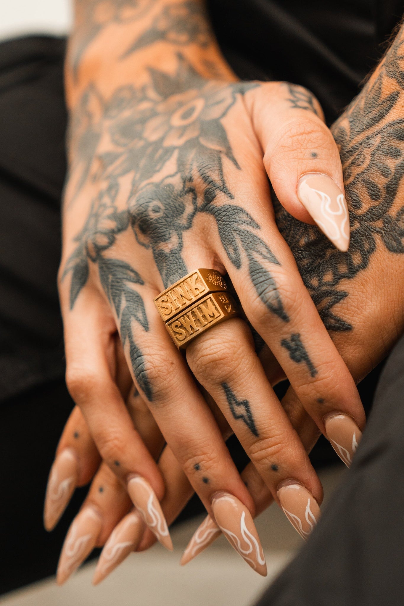 Tattooed hands with long, pointed nude nails featuring white flames display handcrafted rings. The tattoos include floral designs and a lightning bolt, while the stainless steel rings are Relentless Betrayals Gold Sink or Swim Rings.
