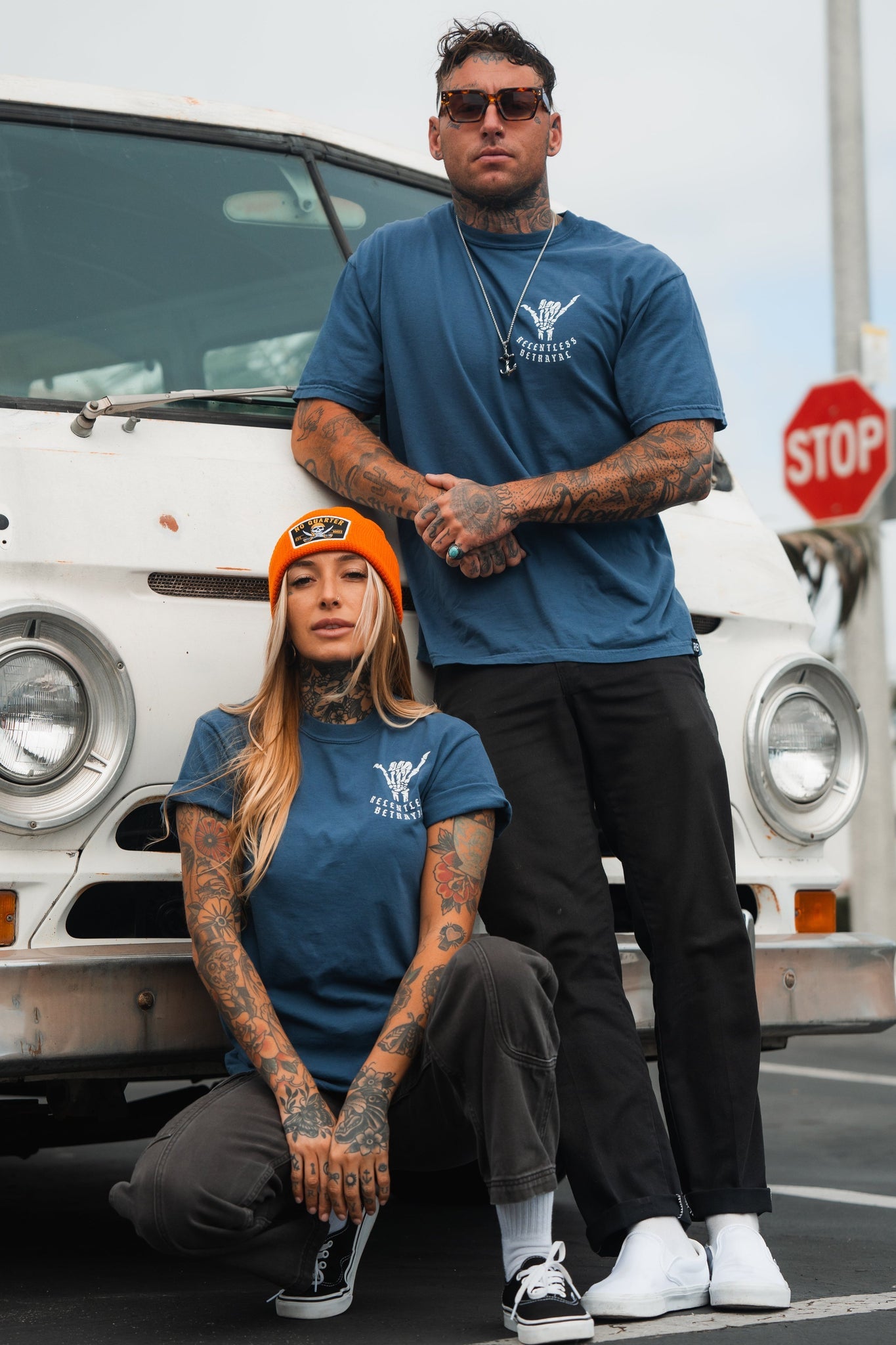 Two tattooed individuals in Sink or Swim Premium Slate T-Shirts by Relentless Betrayal pose in front of a vintage white vehicle, exuding resilience. The person on the right has crossed arms, while the other kneels confidently with an orange beanie. A stop sign is visible behind them.